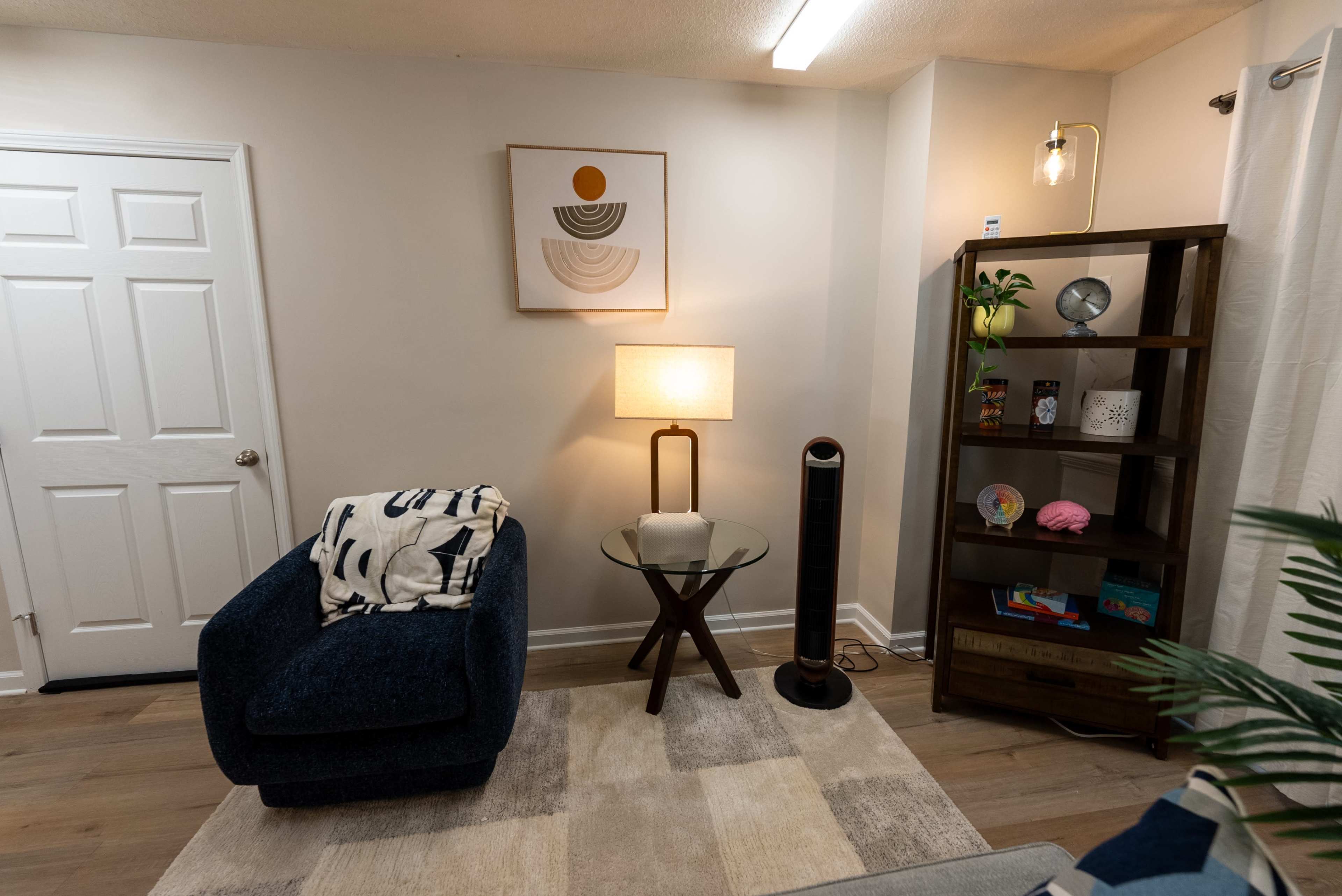The image shows a modern living room with a blue armchair, a round glass table, a lamp, a tall black shelving unit, and a door leading to another room.