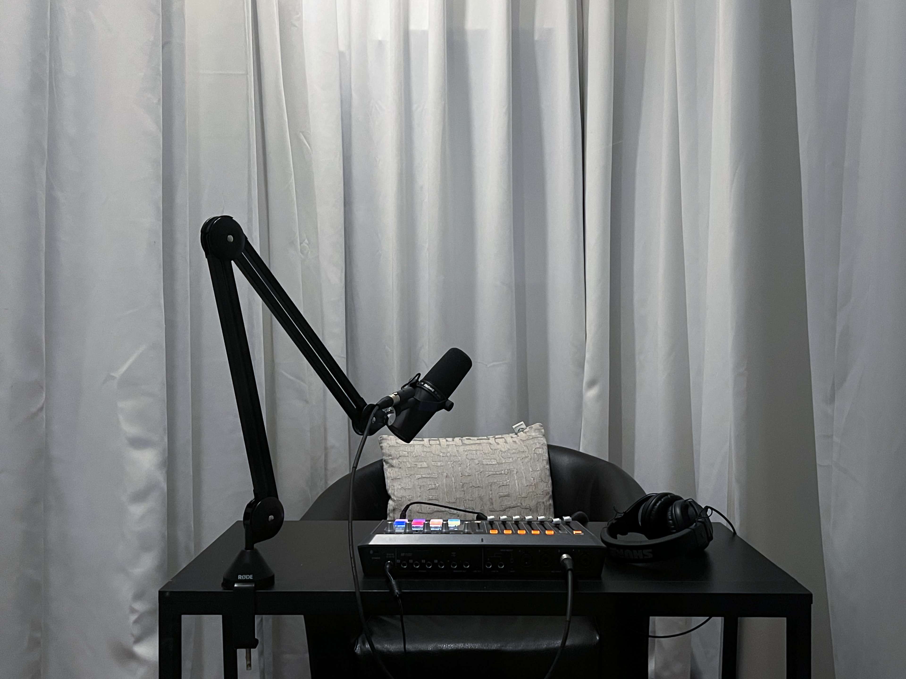 A microphone on a stand is positioned on a black table beside headphones and a sound mixer, with white curtains in the background.