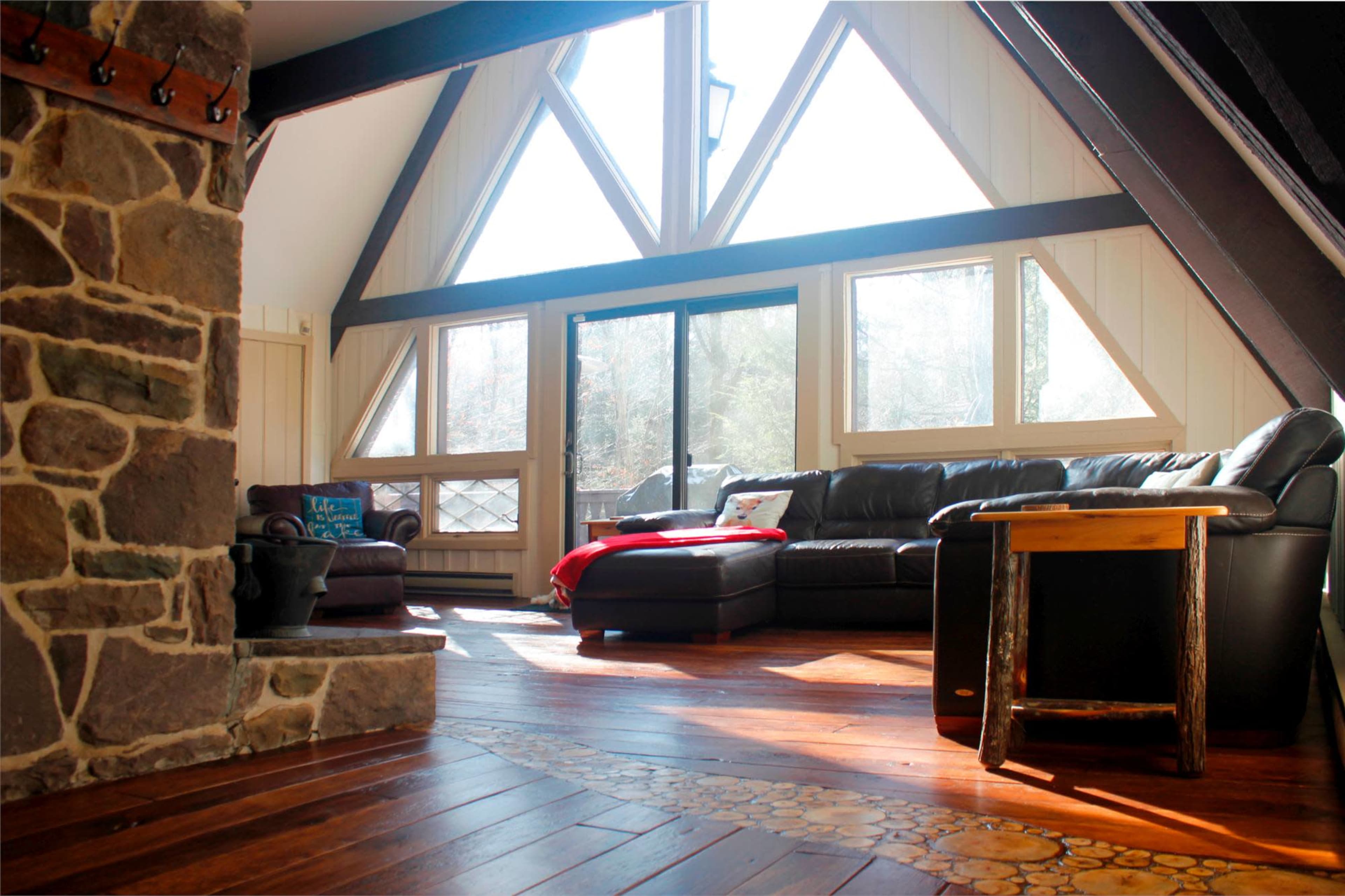 The image shows a cozy living room with large triangular windows, wooden floors, a stone fireplace, and a black leather sofa.