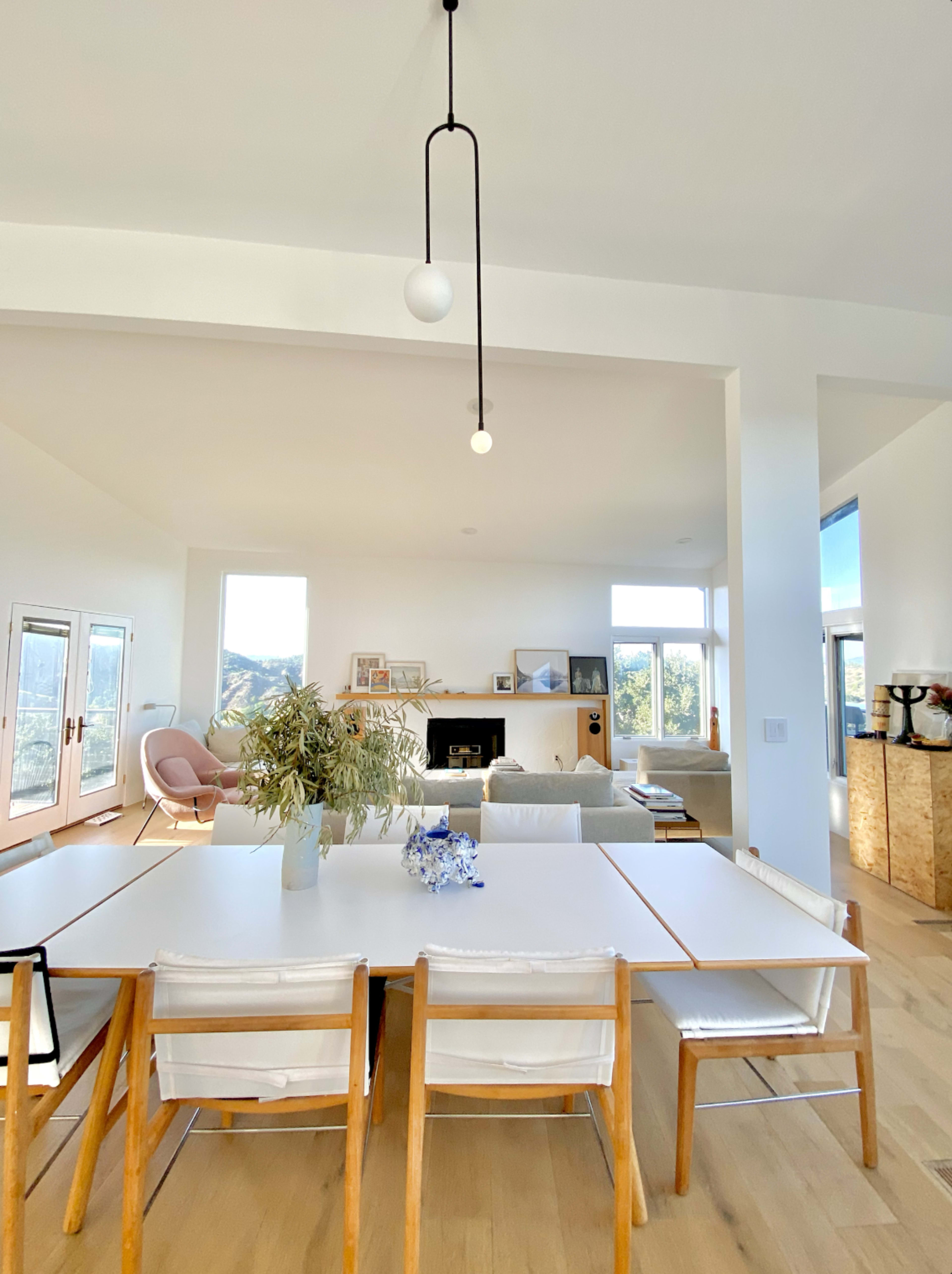 A bright, modern dining area with a large white table and wooden chairs, leading into a spacious living room with a fireplace and large windows.