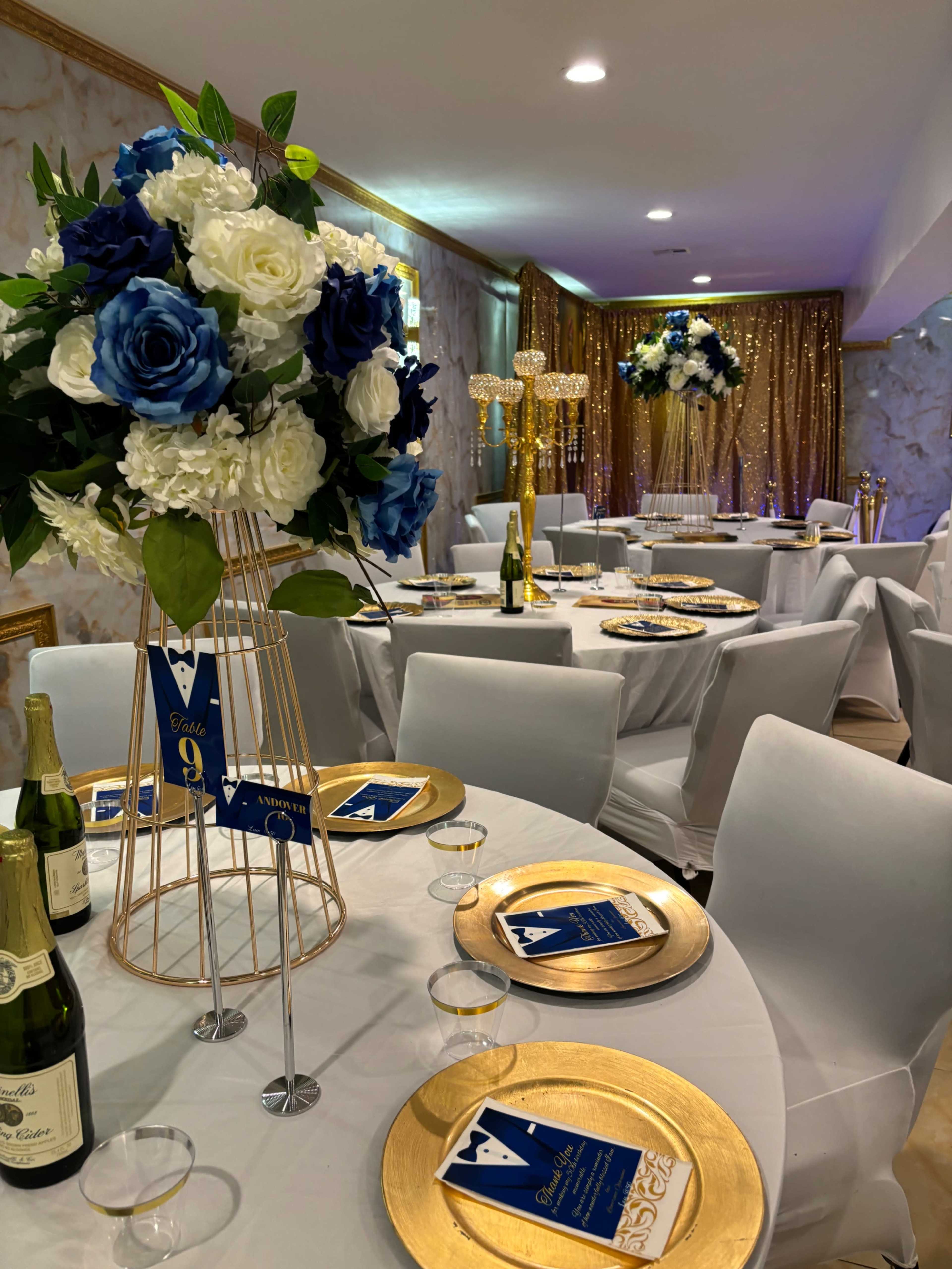 The elegantly arranged dining area features white tables set with gold plates, decorative floral centerpieces, and ambient lighting.