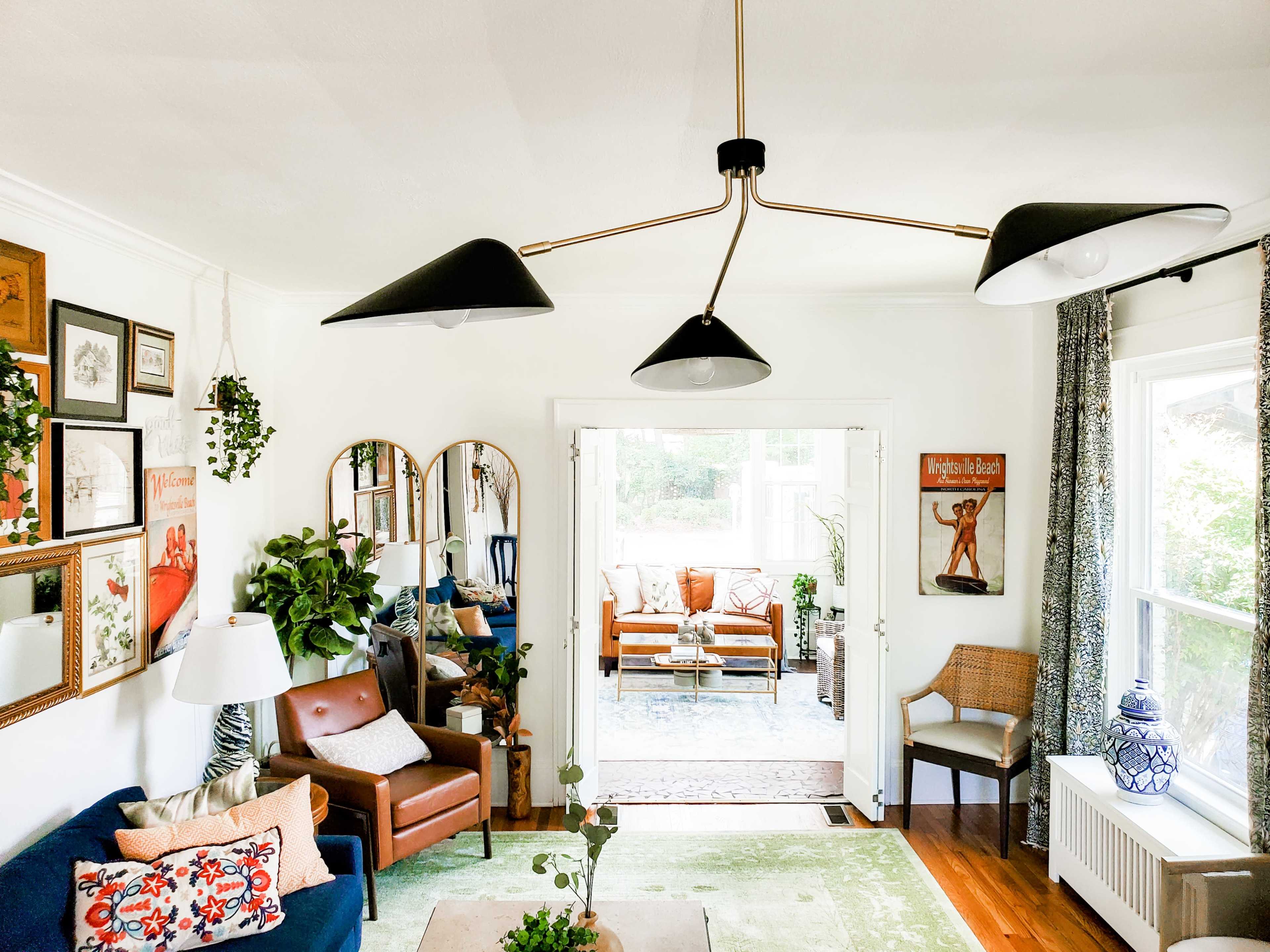 A well-decorated living room features a mix of plants, framed artwork, and a contemporary chandelier, with a cozy seating area and a view into an adjacent sunlit space.