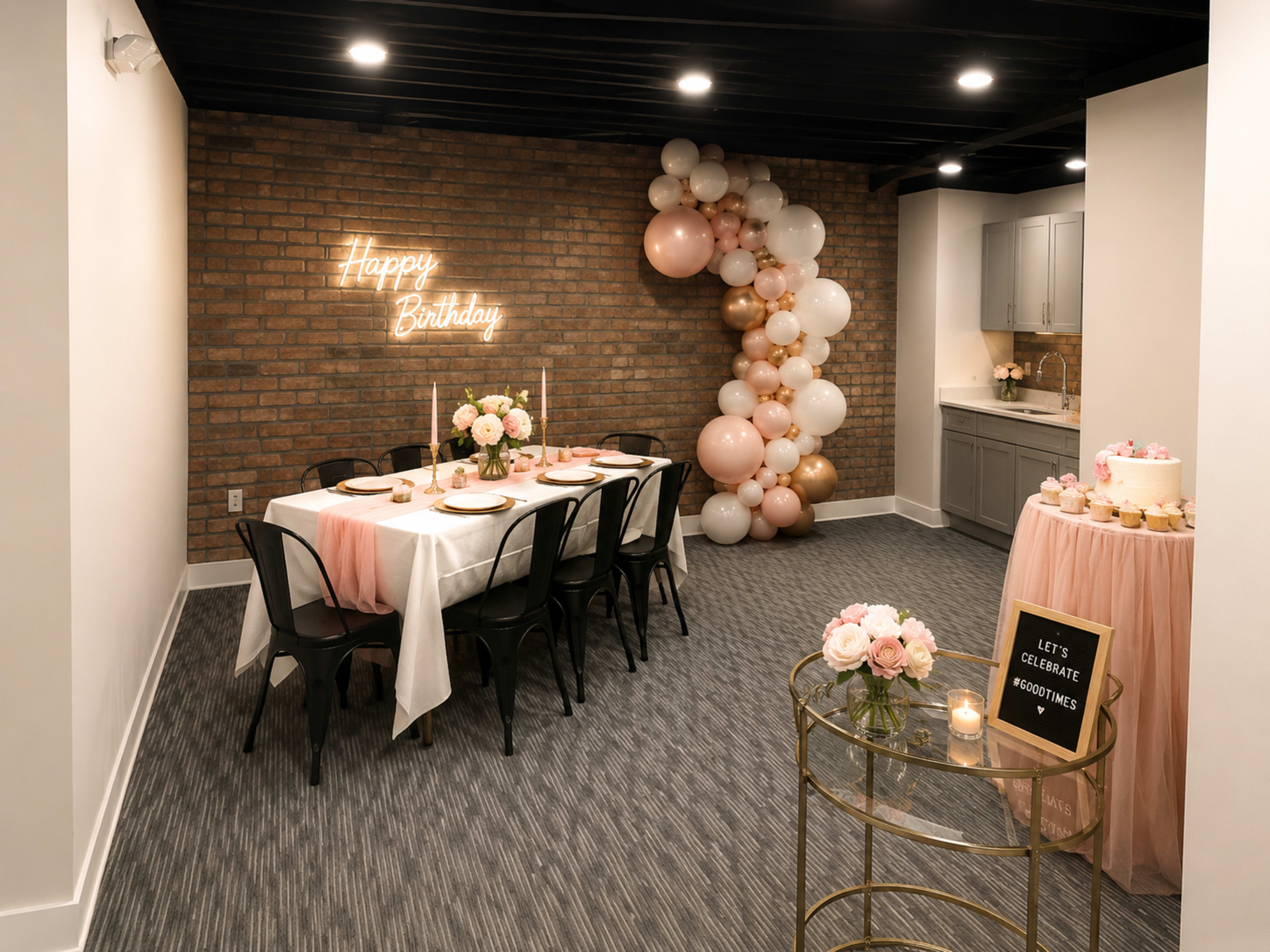 A decorated party room features a long table with white linens, a brick wall with neon "Happy Birthday" signage, and a balloon arch in pastel colors.