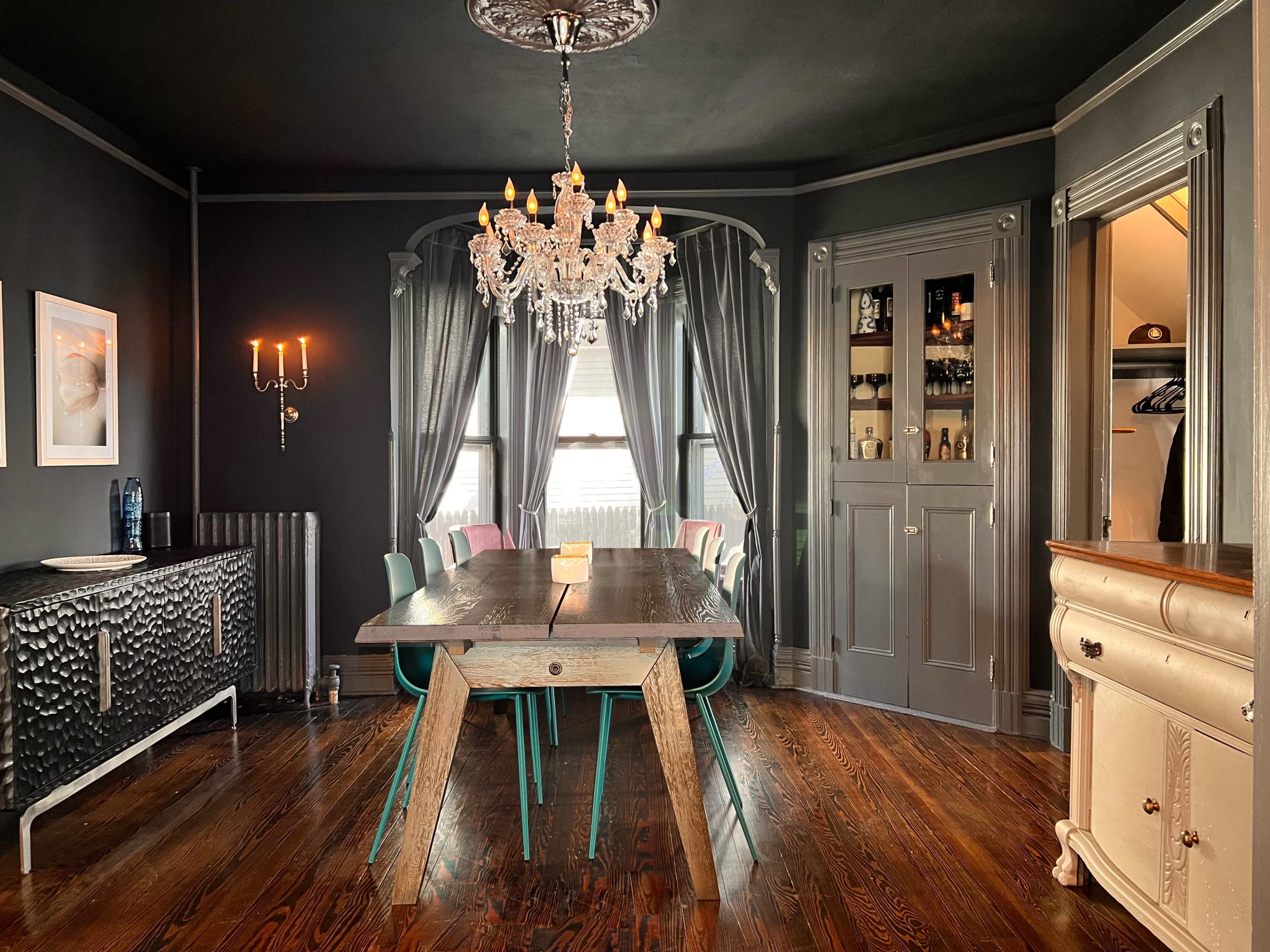 A dining room with a wooden table, colorful chairs, a chandelier, and gray walls, featuring a cabinet and windows draped with curtains.