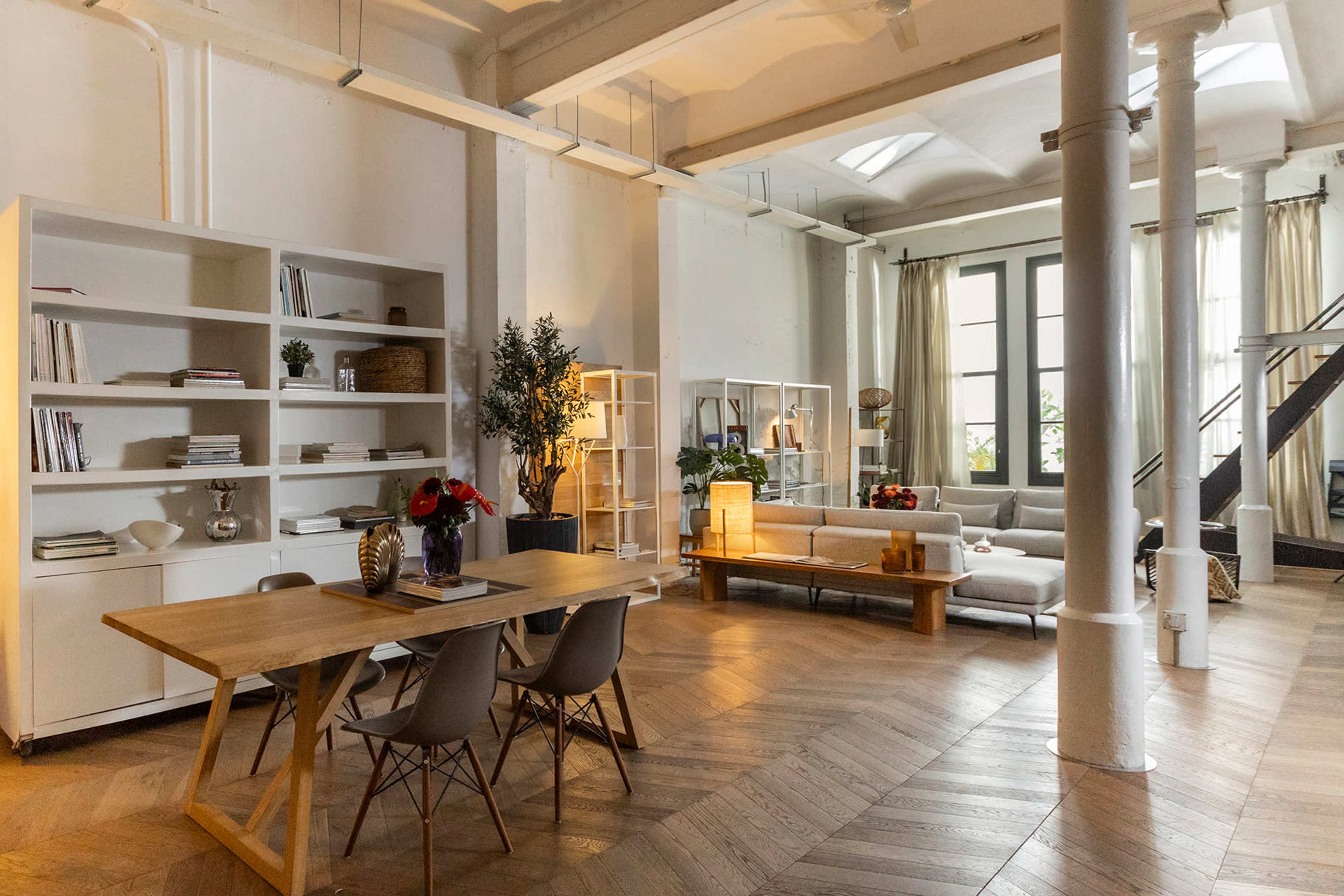 The image shows a spacious, modern living area featuring a wooden dining table, a light-colored seating arrangement, bookshelf units, and large windows that allow natural light to fill the room.