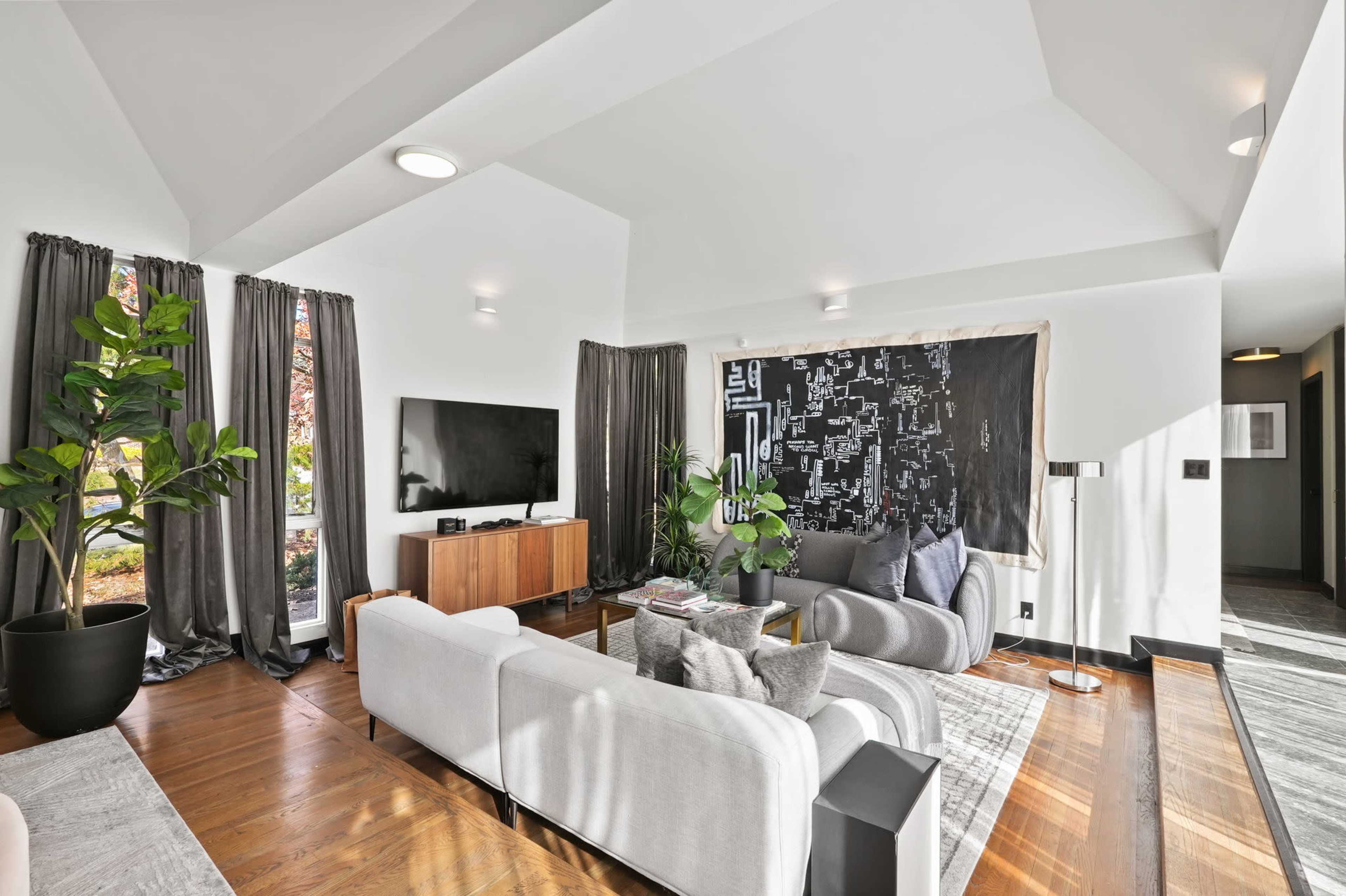 The living room features a modern design with a large television, a wall-mounted artwork, a sectional sofa, and potted plants.
