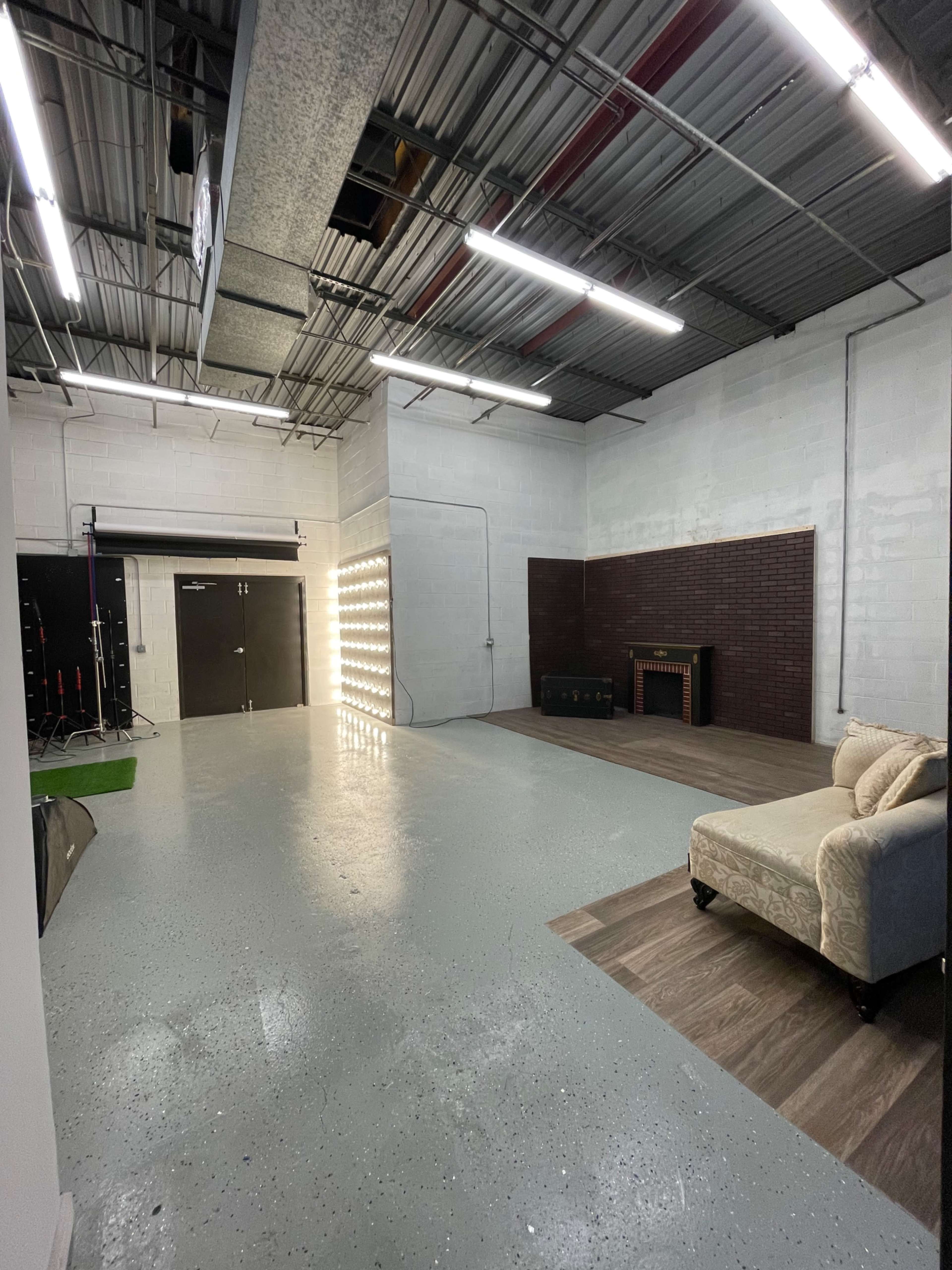 The image shows an empty studio space with a polished concrete floor, a seating area, and a backdrop featuring a wall with lighting fixtures.