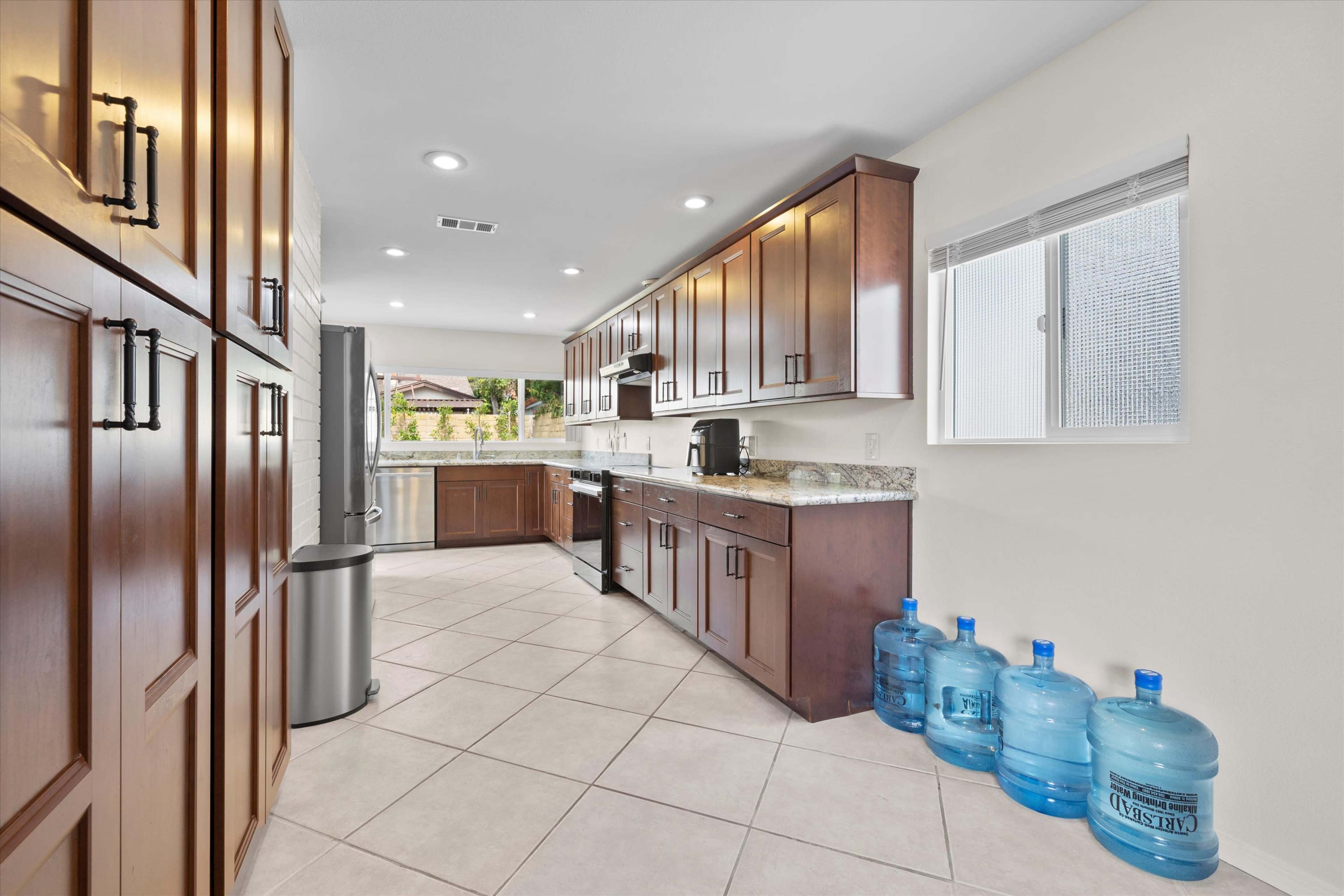 The image shows a modern kitchen with dark wooden cabinets, a stainless steel refrigerator, and several large water bottles lined up against the wall.