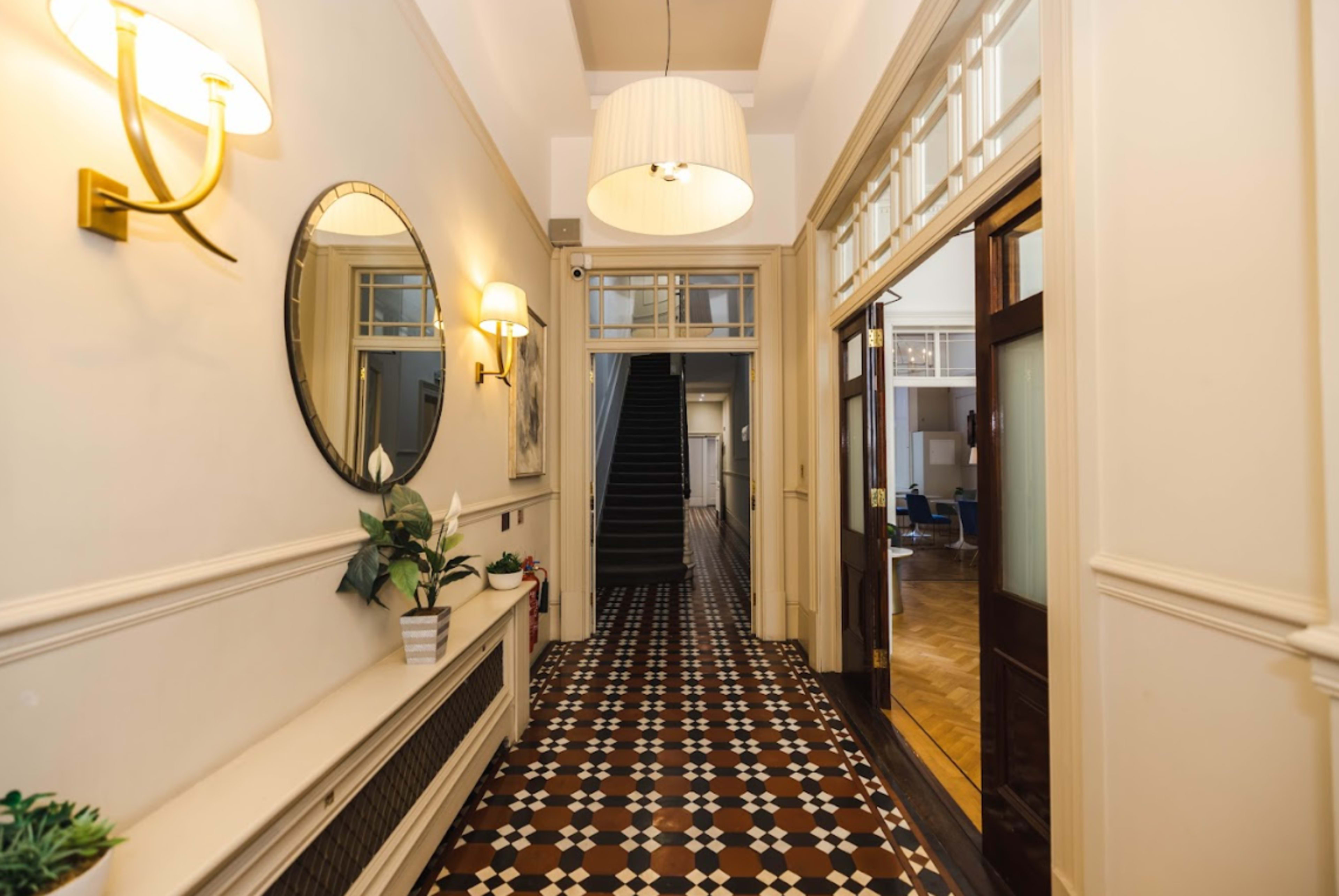 A spacious hallway with a patterned floor, a round mirror, wall sconces, and doors leading to a staircase and additional rooms.