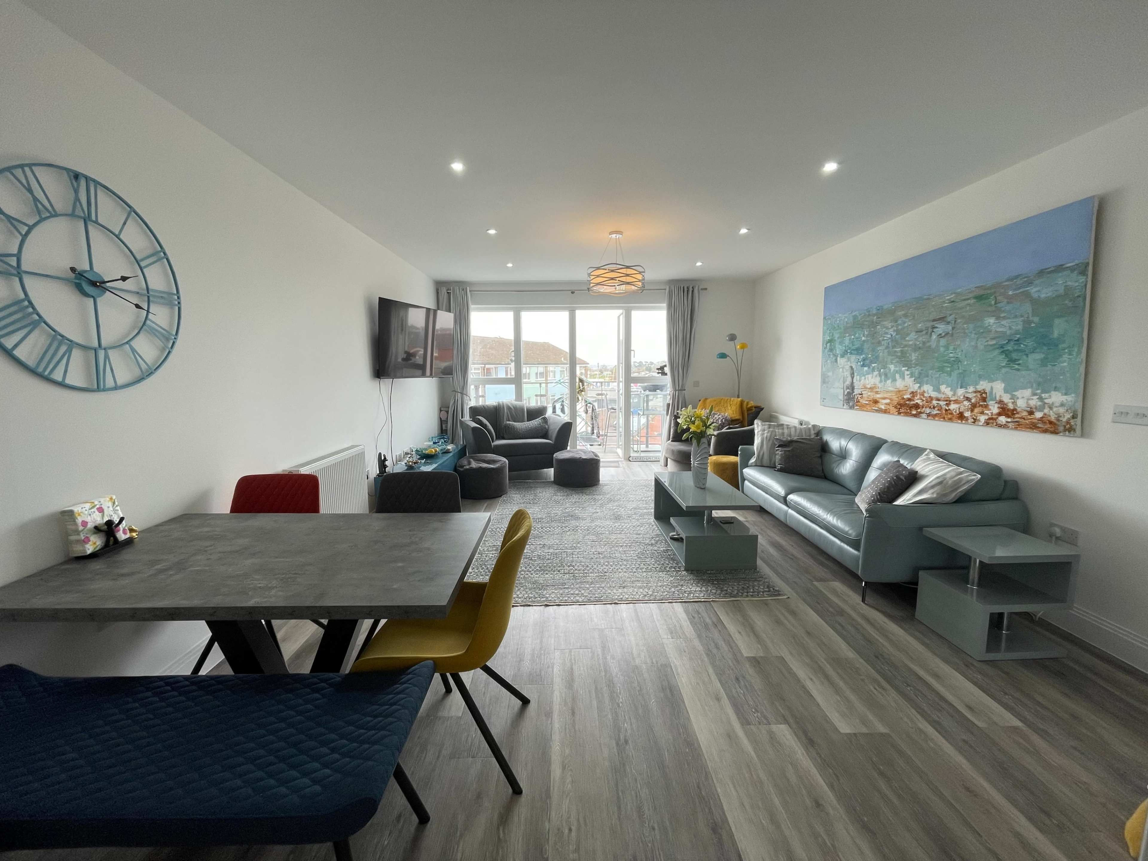 A modern living area features a dining table with colorful chairs, a couch, two armchairs, a television, and large windows opening to a balcony.