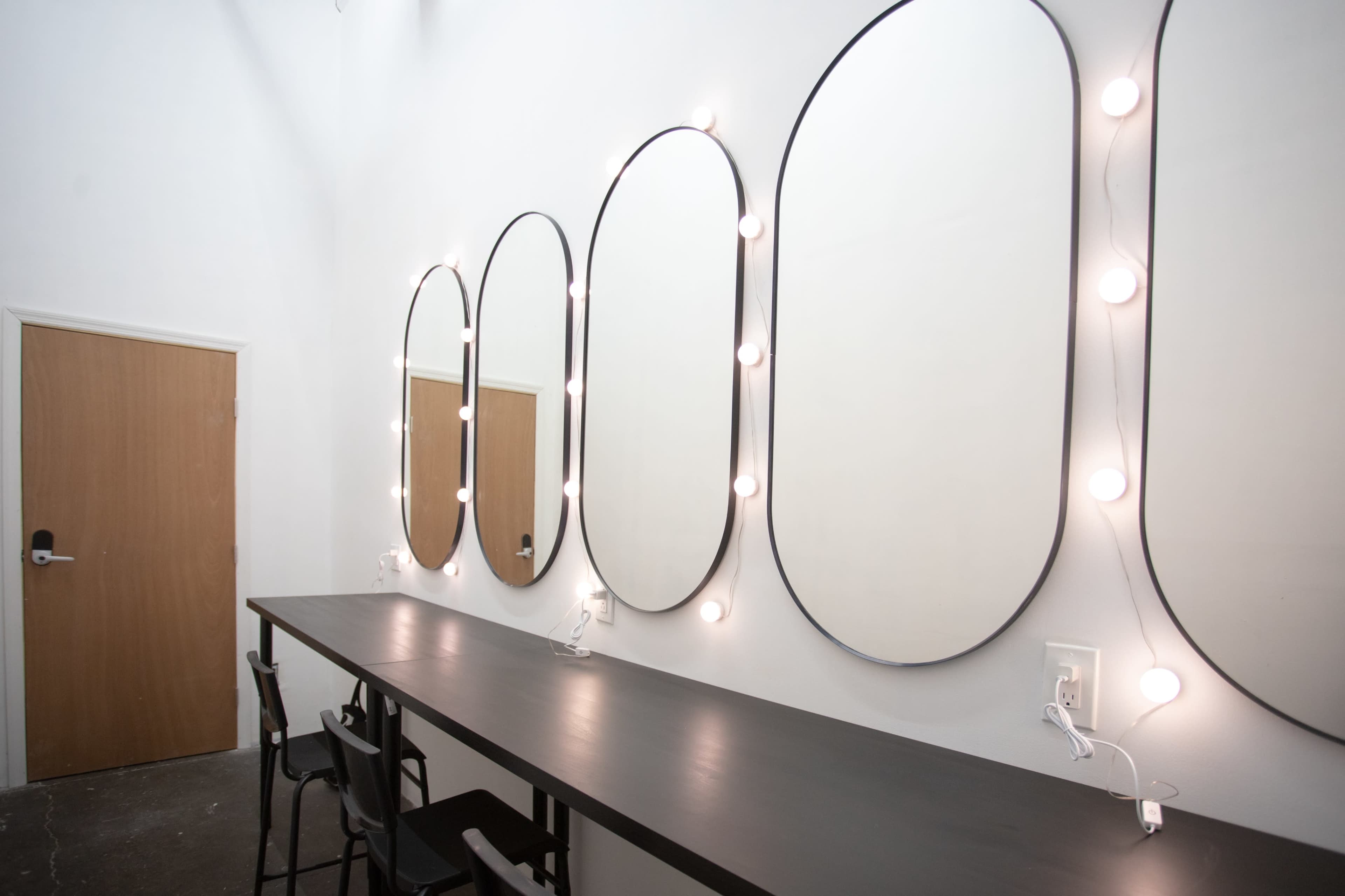 A minimalist vanity area features four oval mirrors with surrounding lights, positioned above a long black counter and a wooden door in the background.