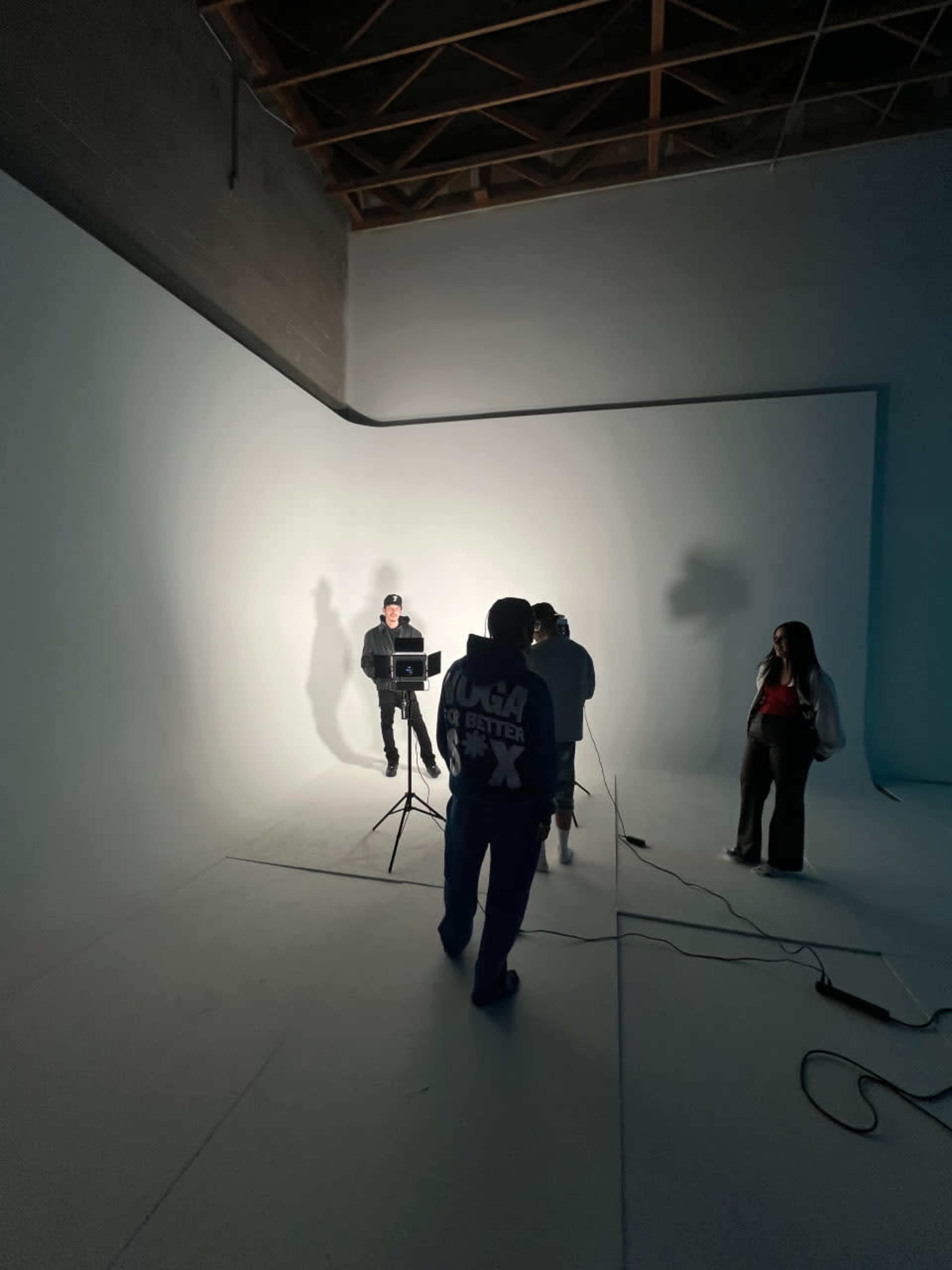 A small group of people is gathered in a well-lit studio with a plain white backdrop, while one person adjusts equipment on a stand.