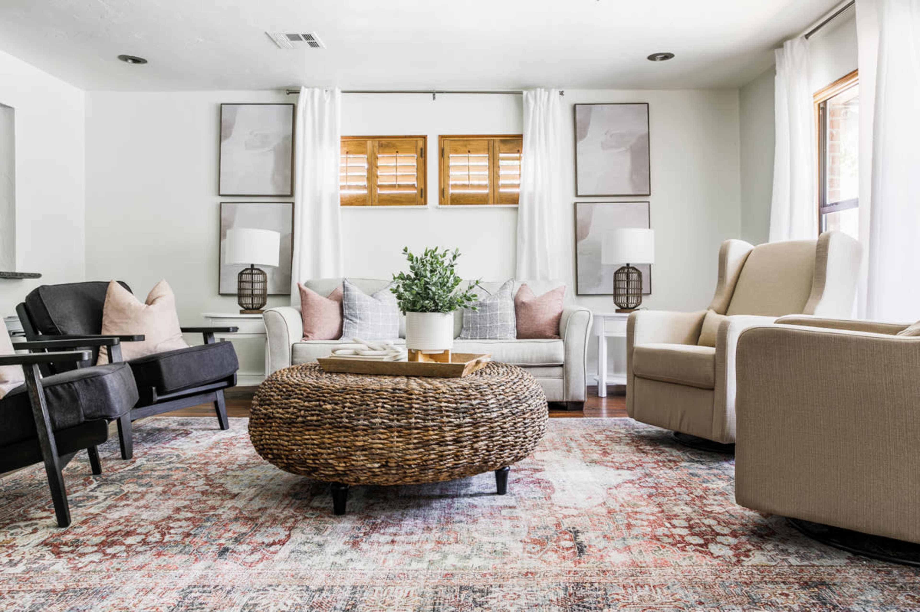 A cozy living room features a round, woven coffee table surrounded by light-colored sofas and accent chairs, with framed artworks on the walls and natural light coming through windows.