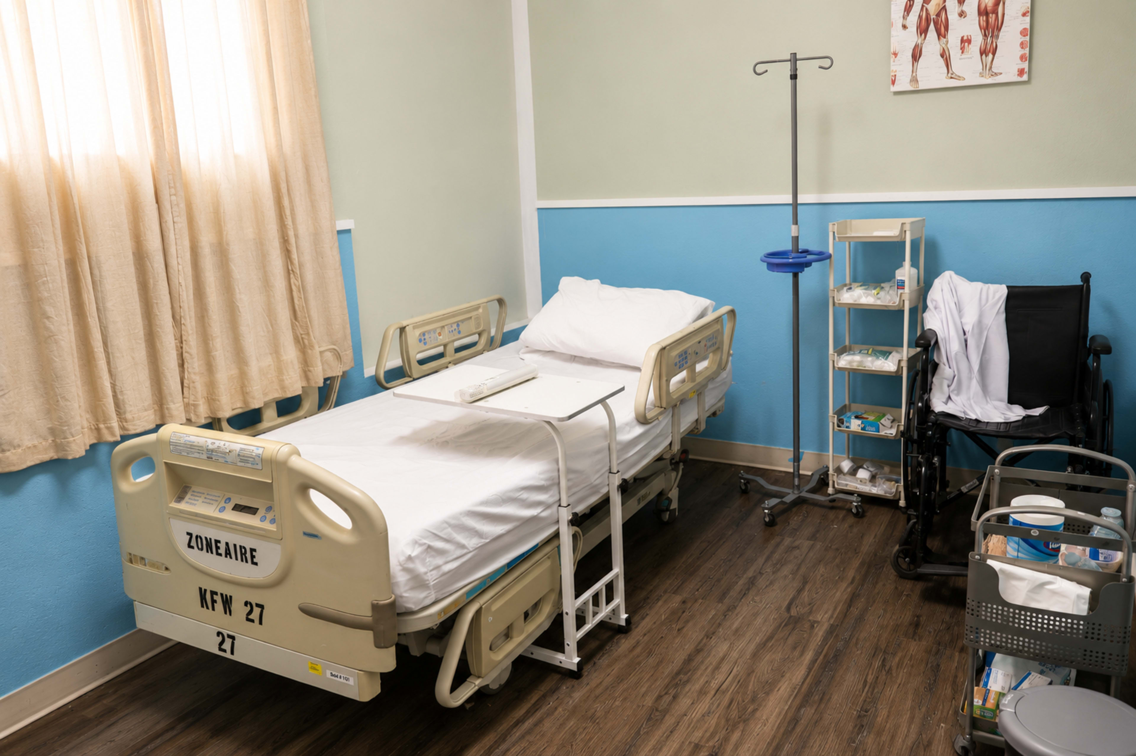The image shows a hospital room with a bed, a wheelchair, a medical cart, and equipment on a wall-mounted stand.