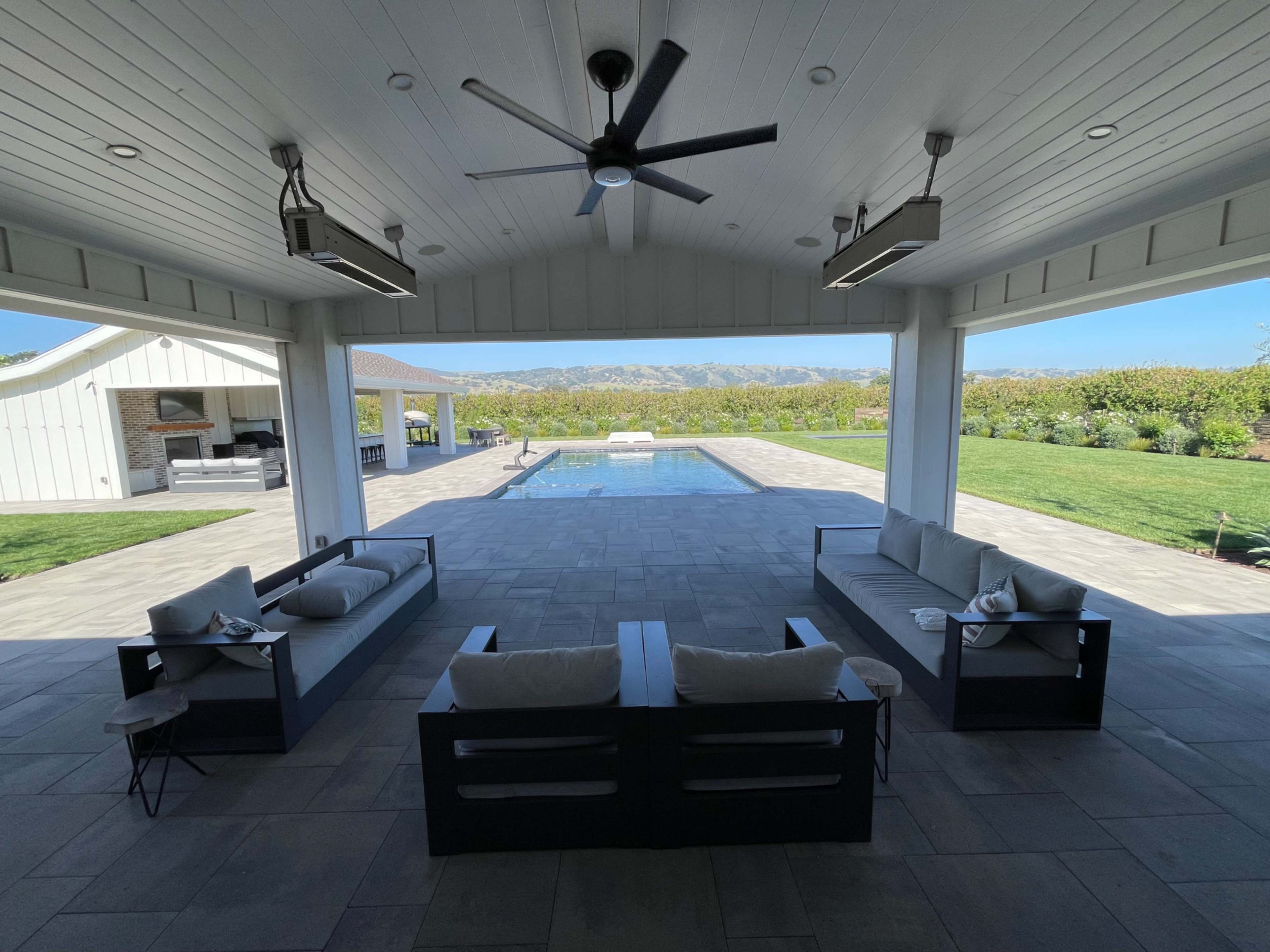 The image shows a spacious outdoor patio with modern seating arrangements overlooking a pool and gardens against a mountainous backdrop.