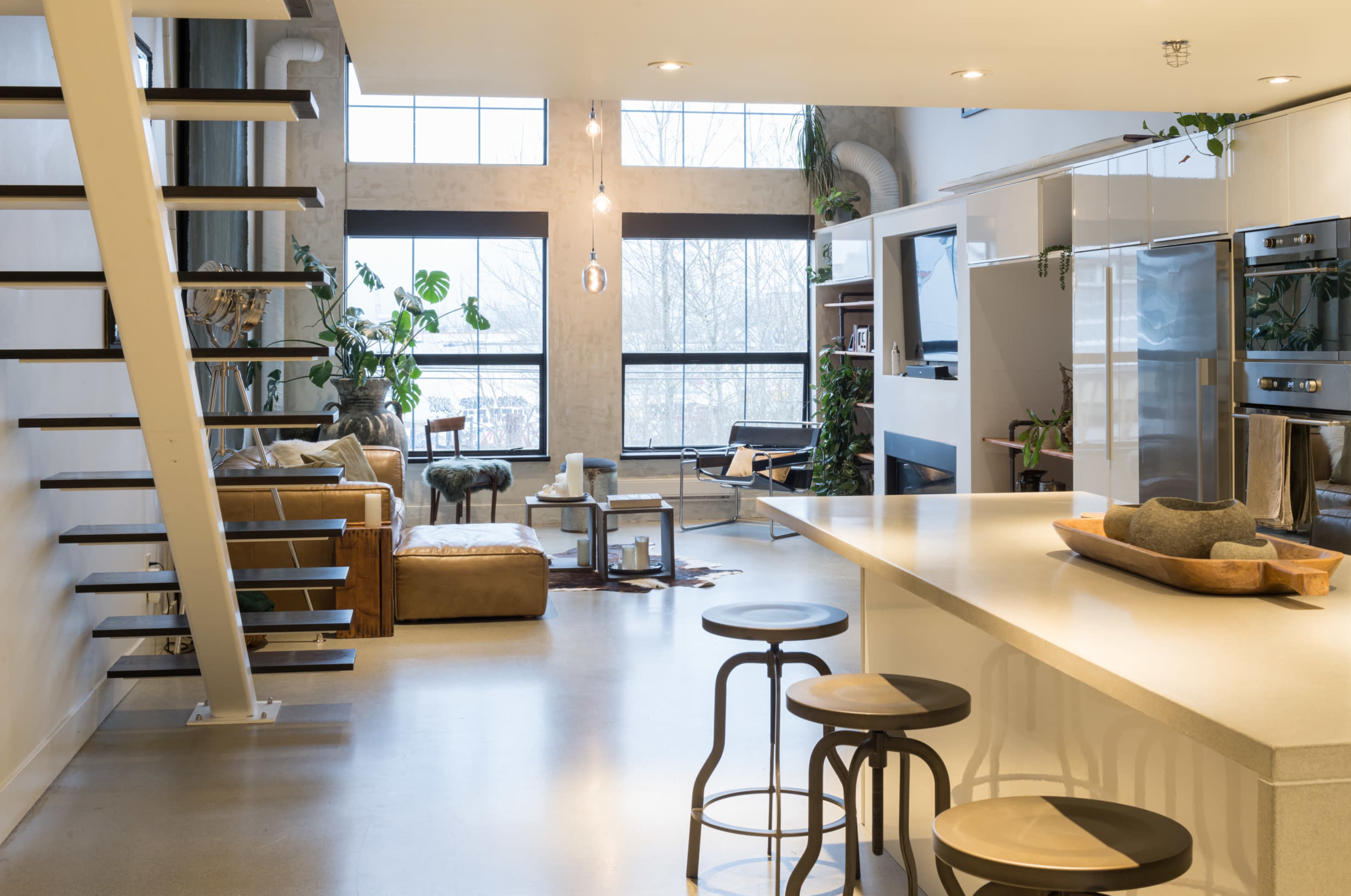 A modern open-concept living space features a kitchen with a central island, bar stools, and a staircase leading to an upper level, while a cozy seating area is visible through large windows filled with plants.