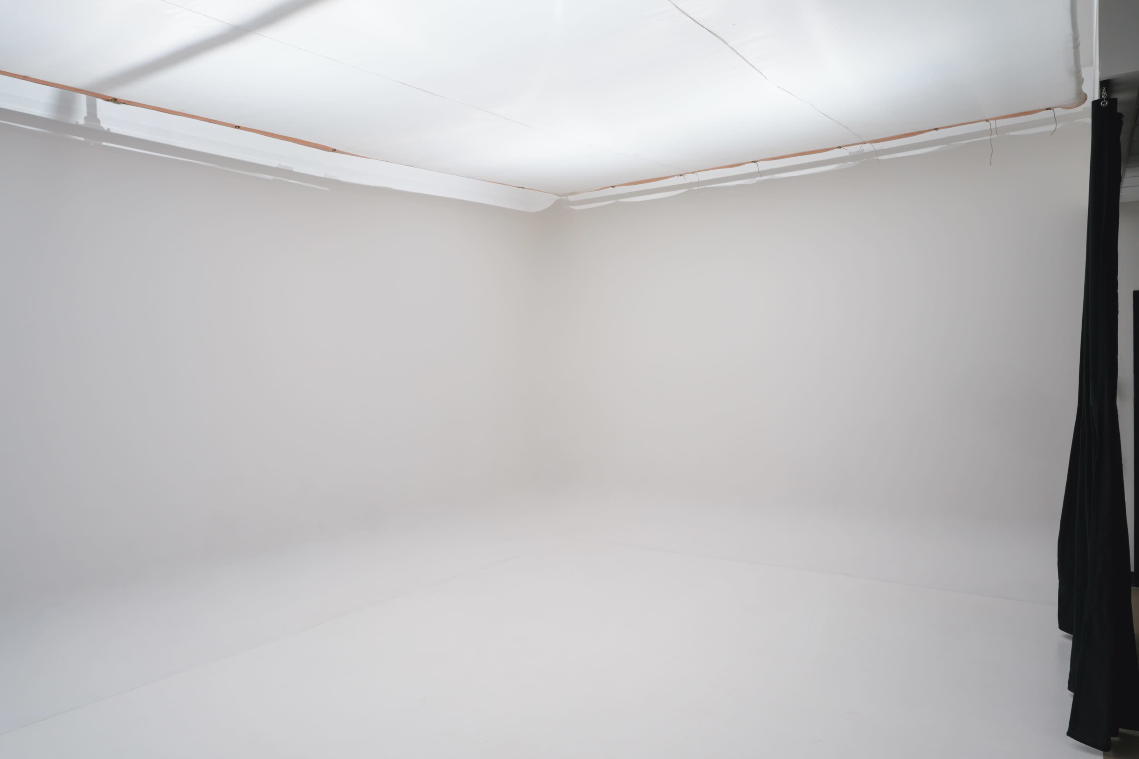 The image shows a blank, well-lit studio space with white walls and floor, featuring a black curtain on one side.
