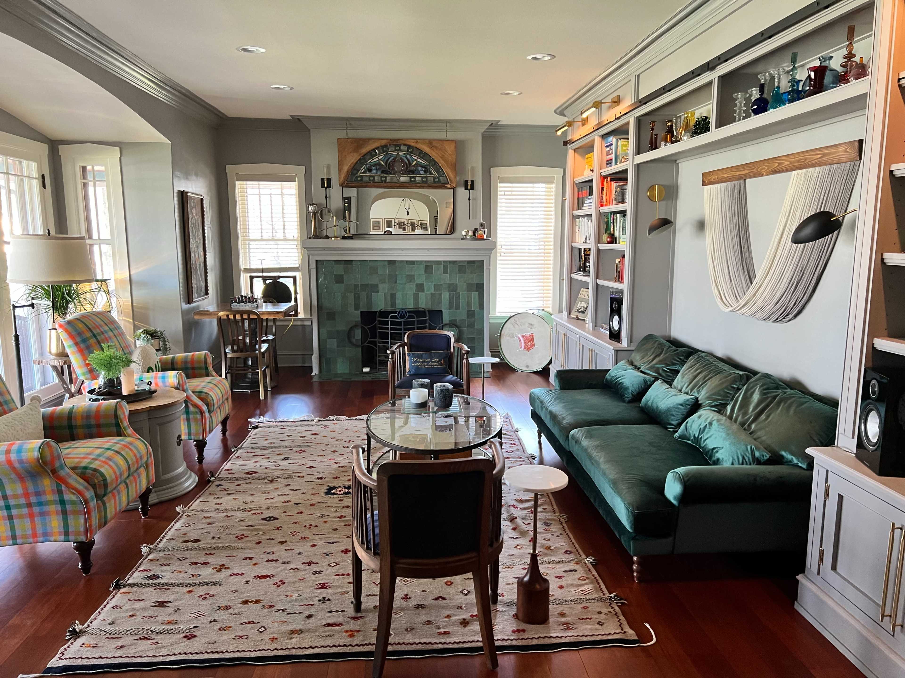 The living room features a green sofa, a patterned armchair, and a decorative fireplace, with shelves displaying various items and a large area rug on the wooden floor.