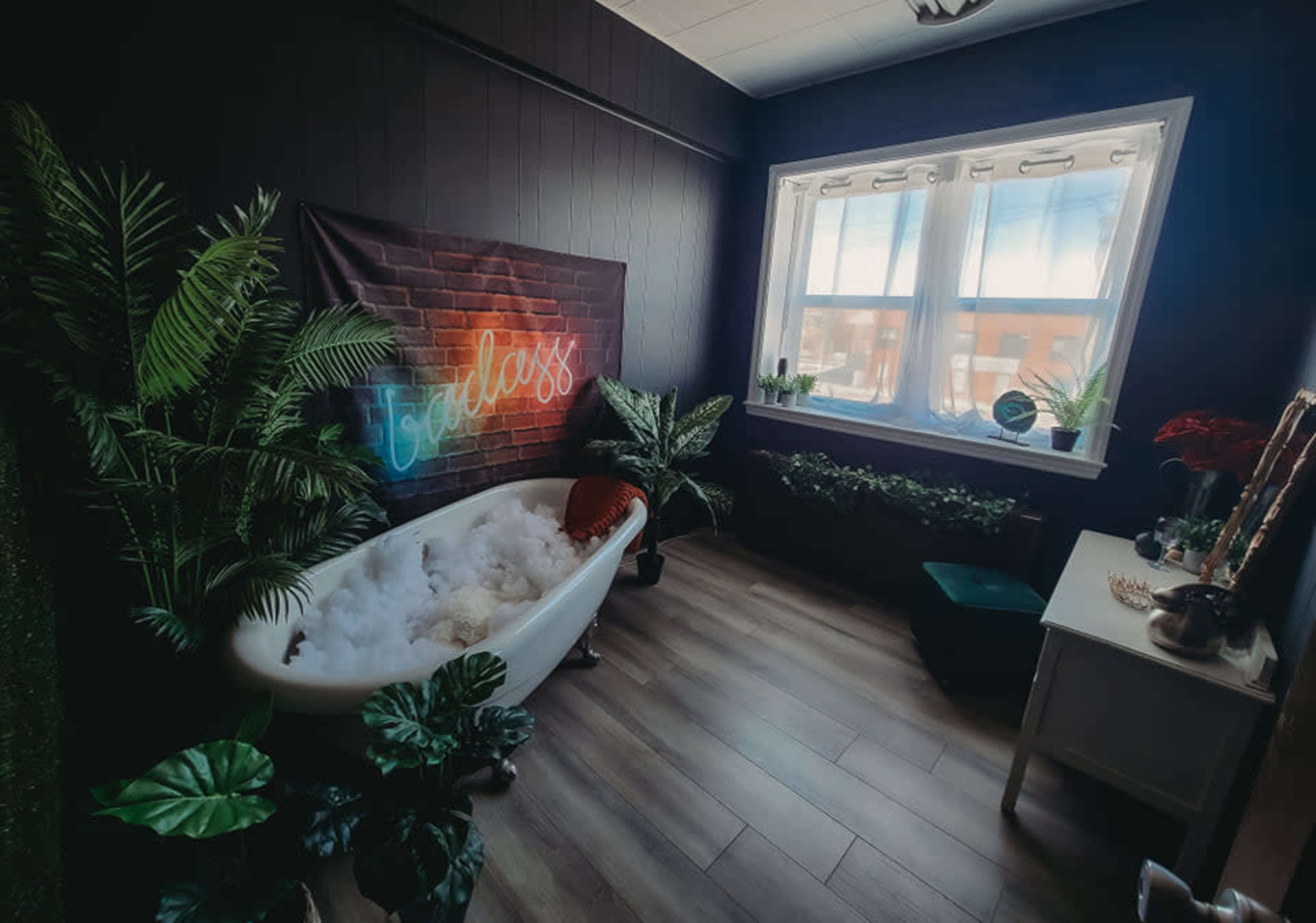 A small, stylish bathroom with a vintage bathtub filled with bubbles, surrounded by indoor plants and a large window letting in natural light.