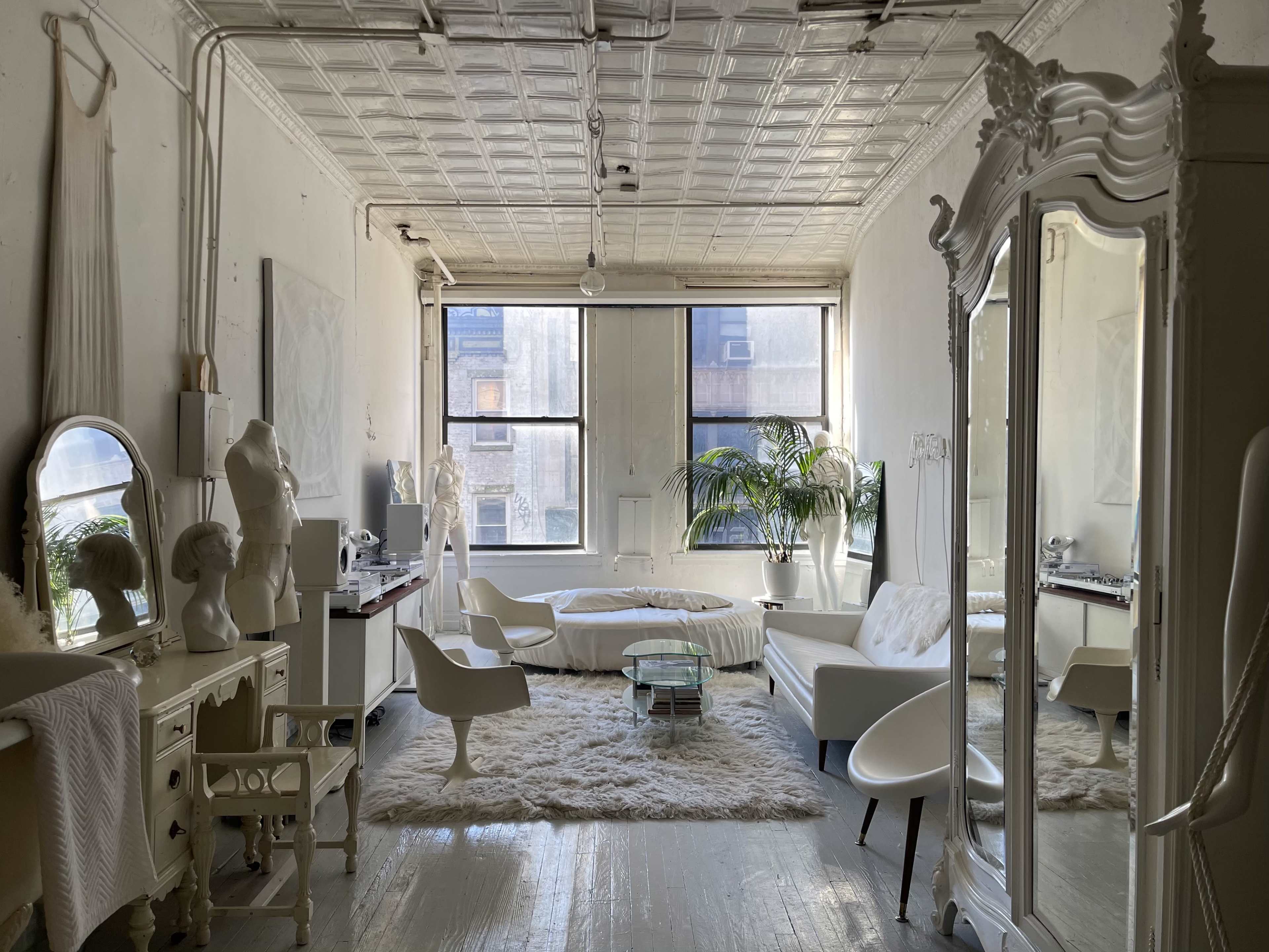 The image shows a bright, minimalist living room with white walls, a plush rug, modern furniture, and large windows allowing natural light to enter.