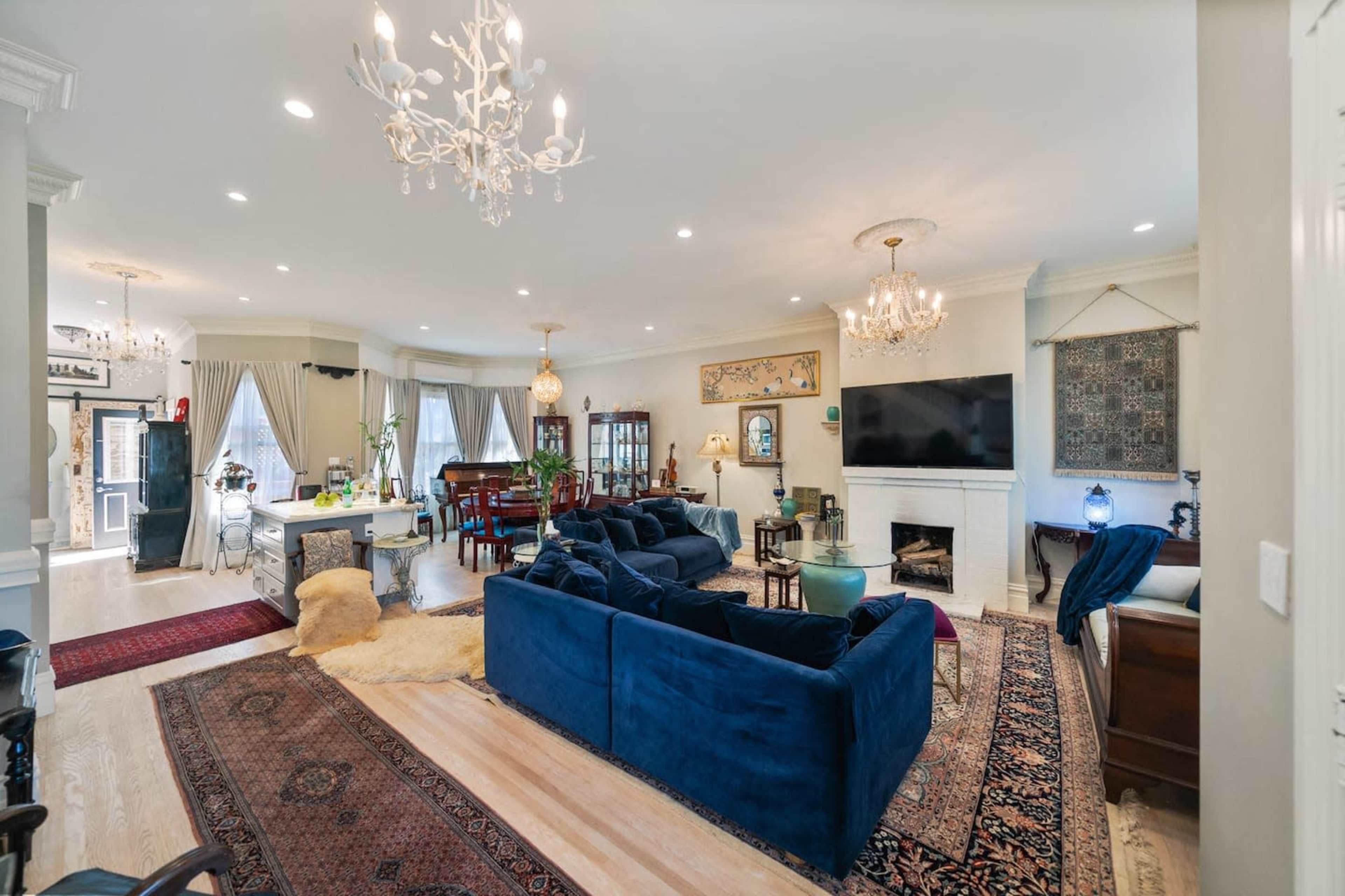 A spacious living room featuring a blue velvet sectional sofa, a large television mounted above a white fireplace, and various decorative elements including chandeliers and rugs.