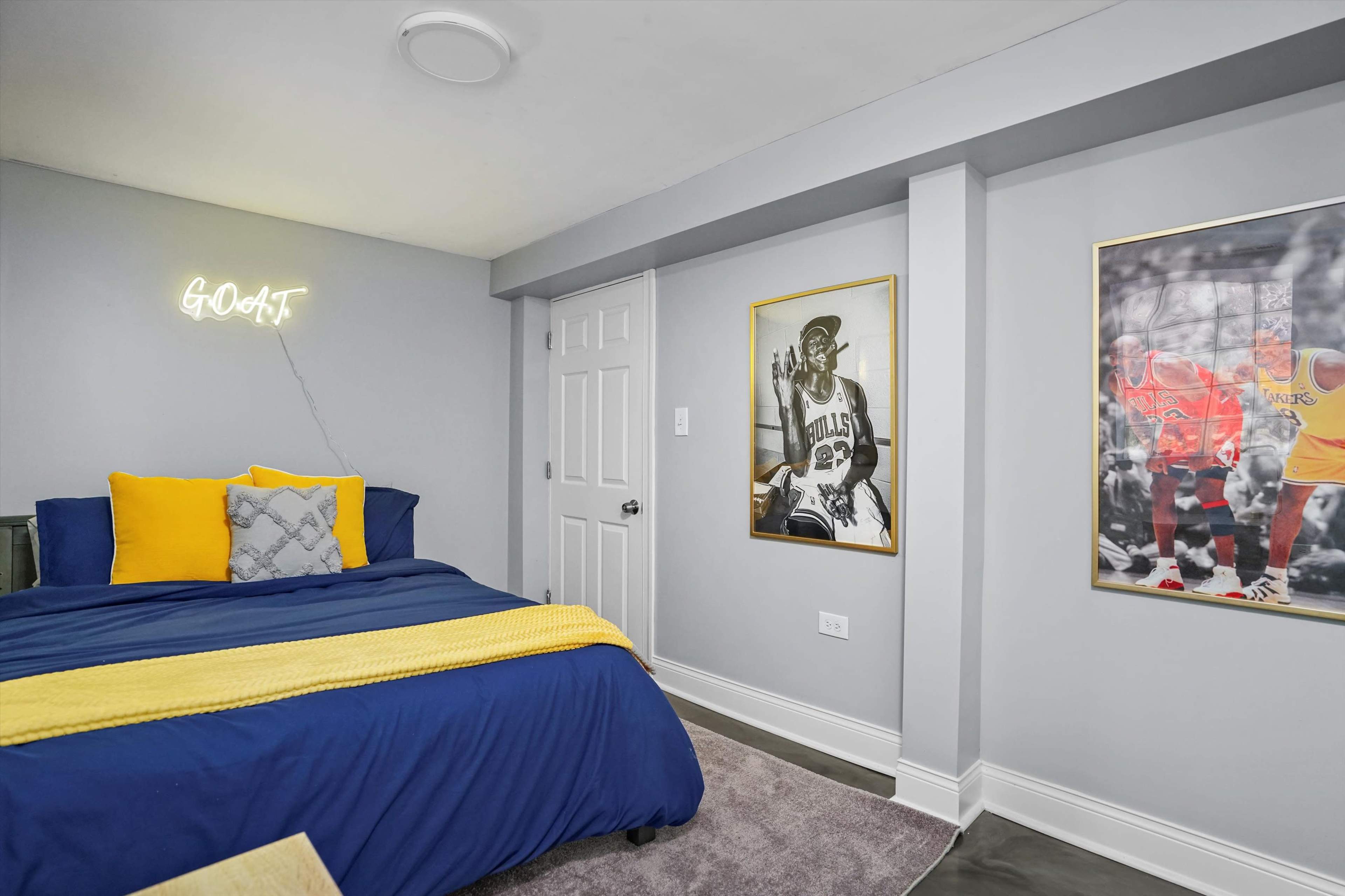 The image shows a bedroom with a blue bedspread, yellow decorative pillows, and framed sports posters on the walls.