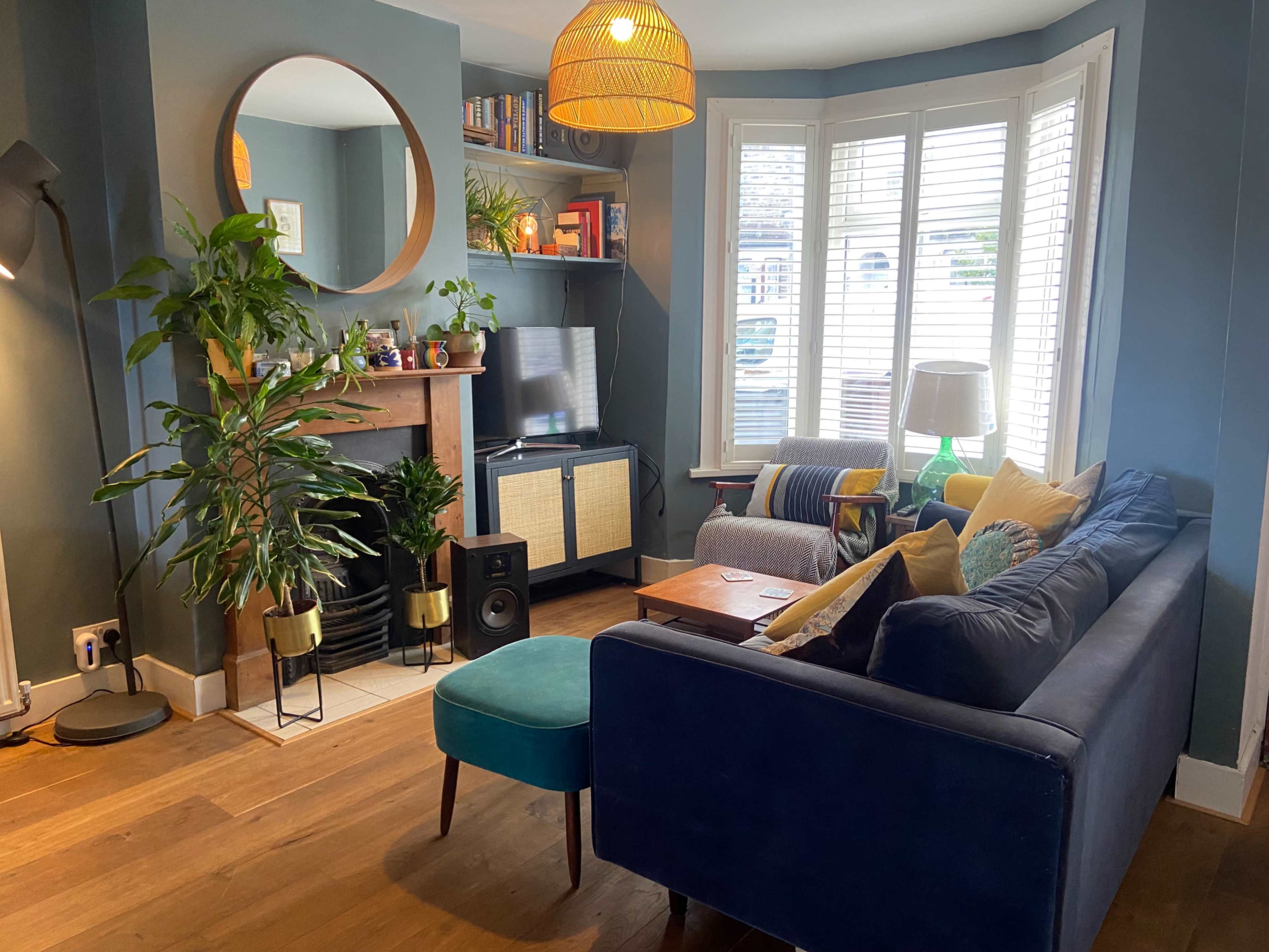 A cozy living room with a blue sofa, a small green ottoman, and a wooden coffee table, surrounded by plants and a fireplace.