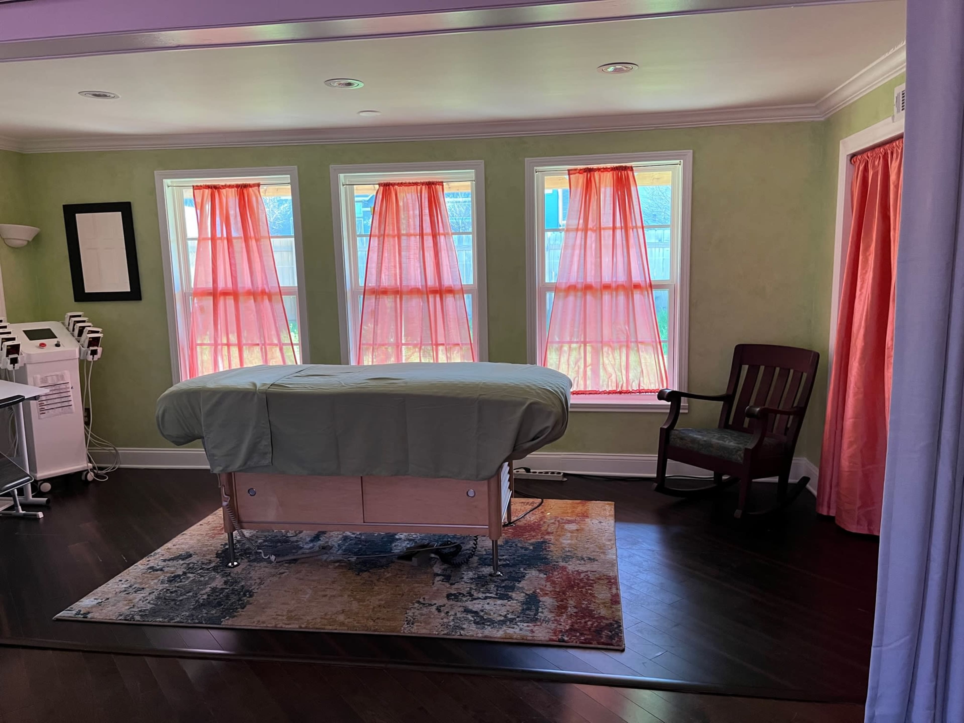 A therapy room with a massage table covered by a green sheet, surrounded by windows with flowing orange curtains and a wooden chair in the corner.