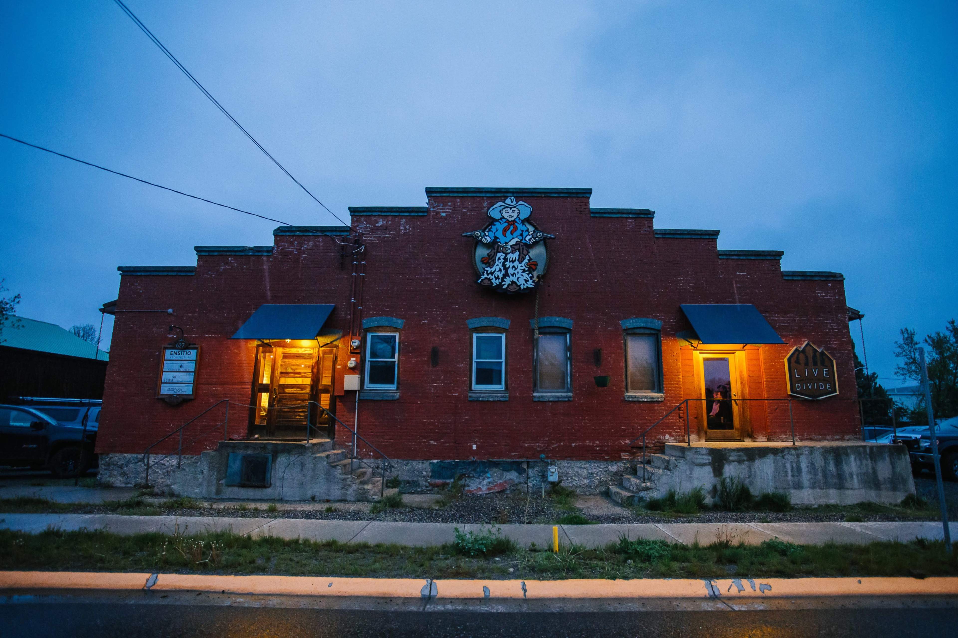 A brick building with a decorative mural on the facade, illuminated by warm lights on either side of the entrance, set against an evening sky.