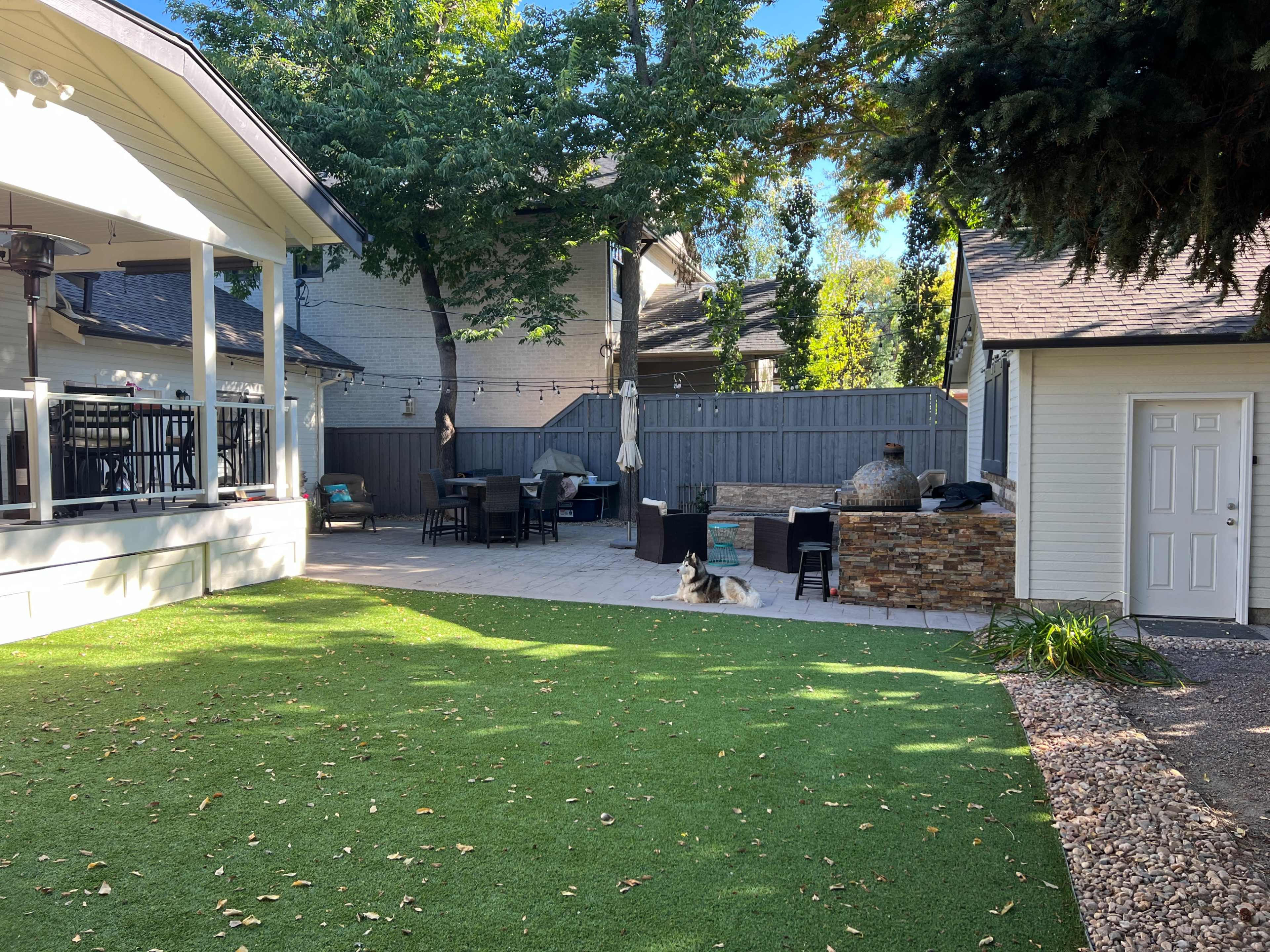 The image shows a backyard with a patio area featuring a dining set, a stone fire pit, and a green lawn, along with a small building and a dog resting on the grass.