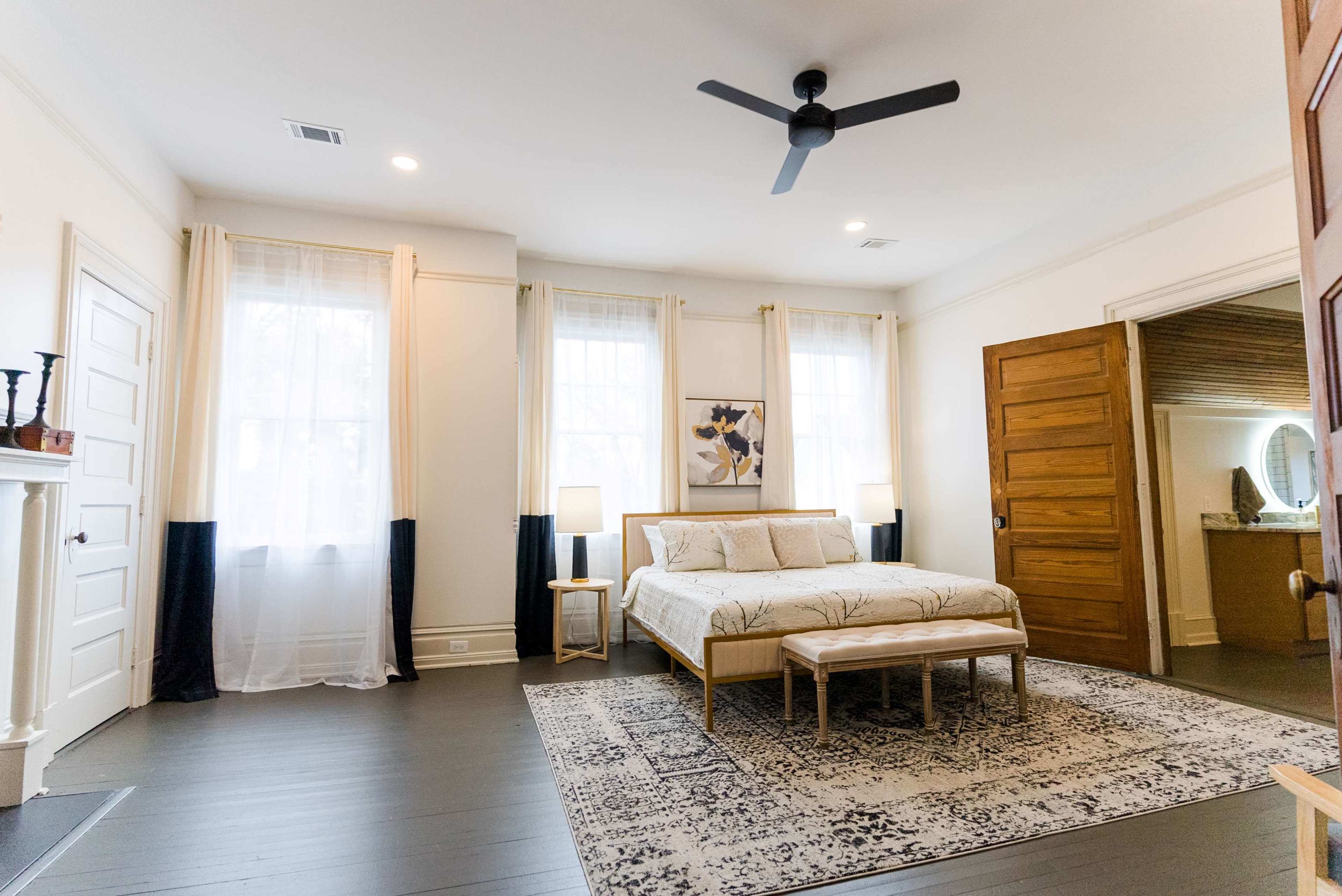 A spacious bedroom features a central bed with a patterned duvet, two bedside lamps, and a decorative area rug on a dark floor.