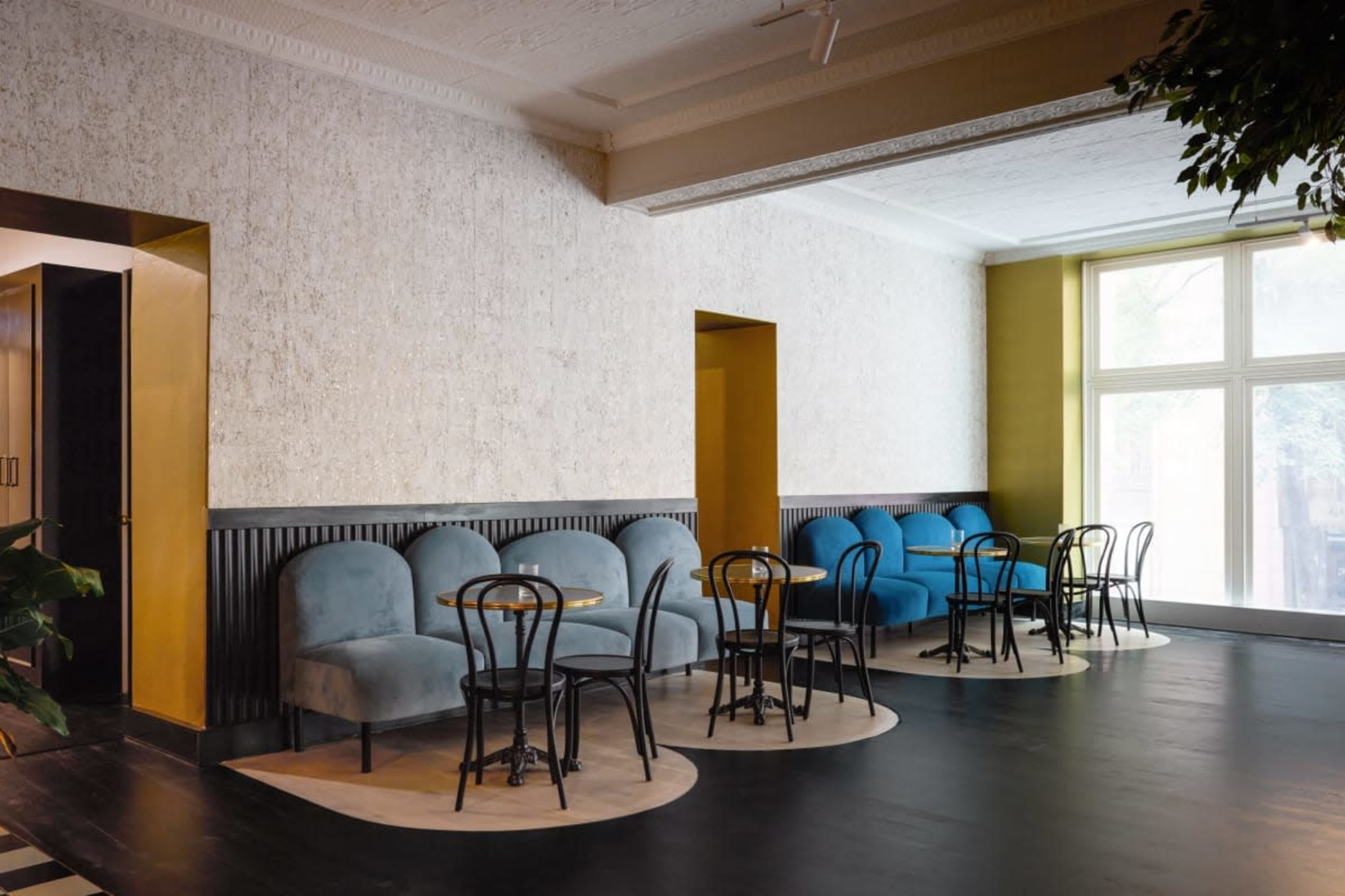 A modern café interior with blue and beige upholstered seating, round tables, and large windows allowing natural light to enter.