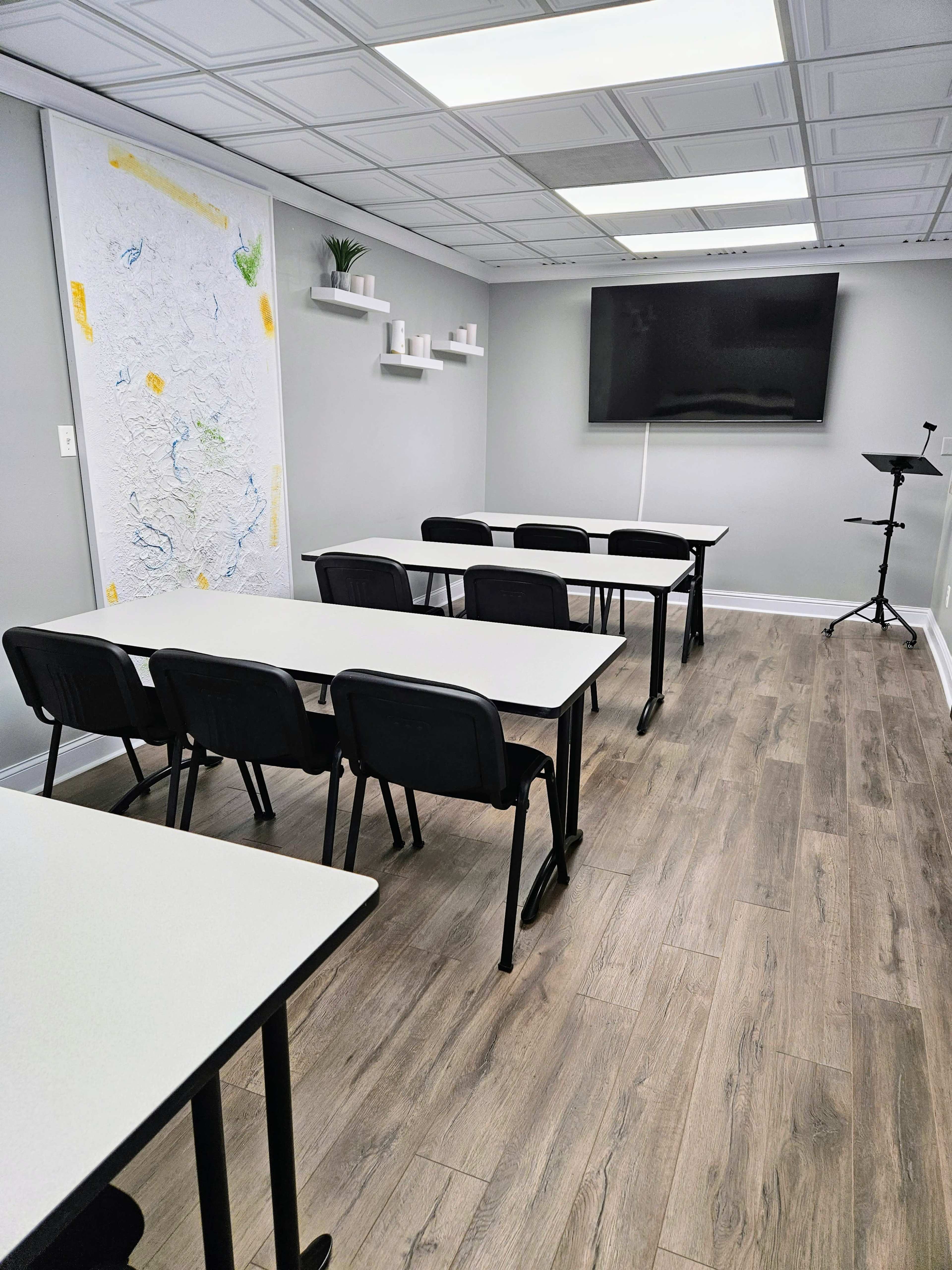 A modern classroom or meeting room with several tables and chairs arranged in rows, a large screen on the wall, and a whiteboard with a map sketch.