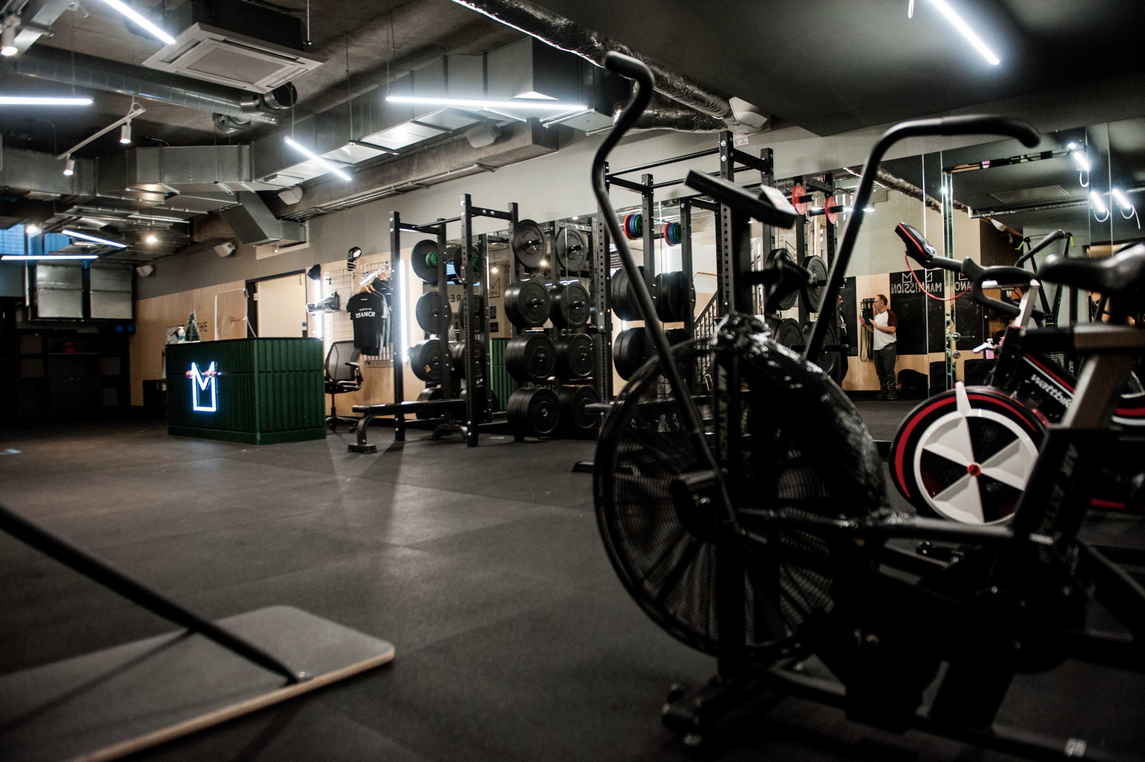 The image shows a modern gym interior with various exercise equipment, including stationary bikes and weightlifting machines.