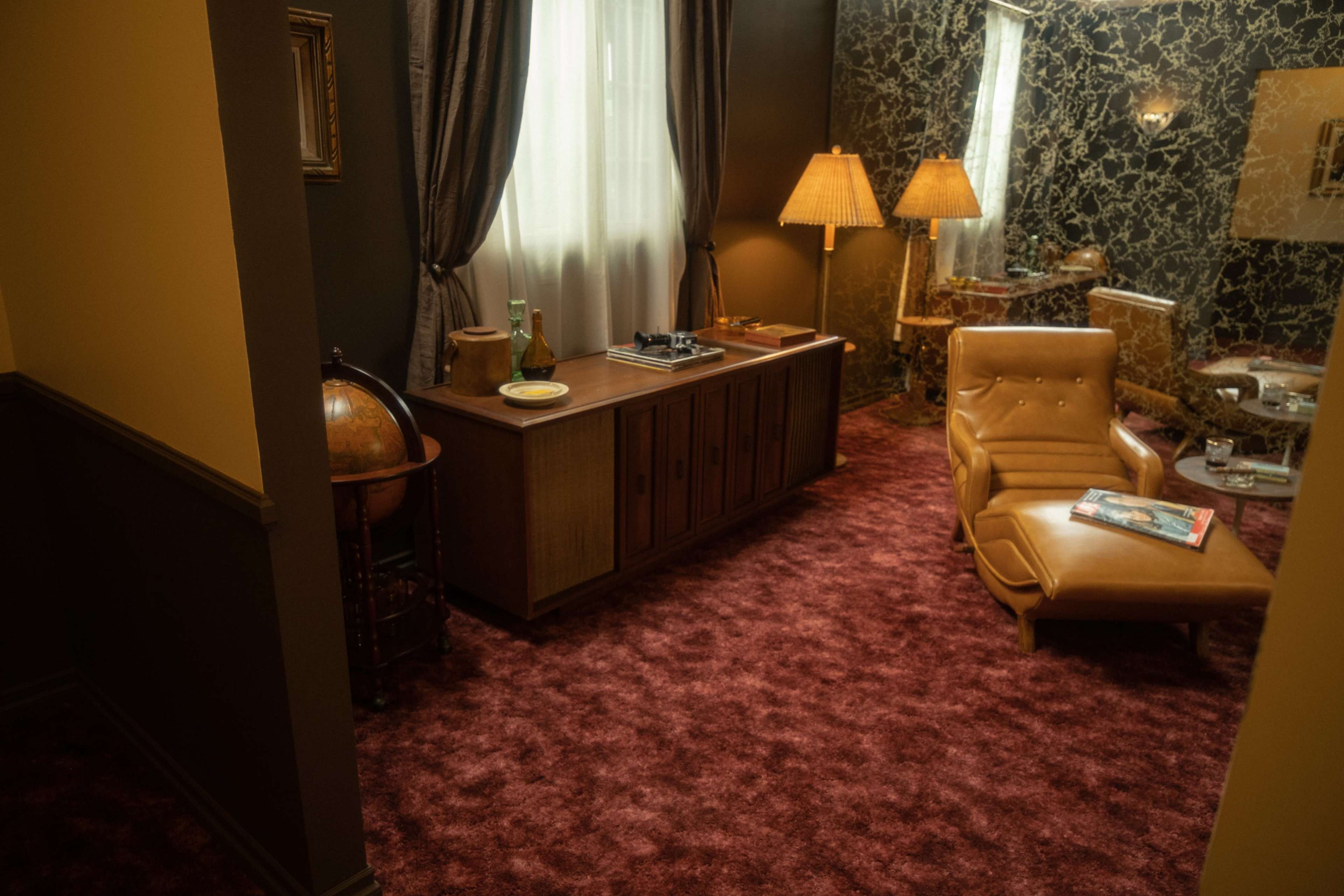 The scene features a room with a vintage decor, including a reclining chair, a wooden cabinet, a retro globe, and two lamps, all set against a patterned wall and plush carpet.