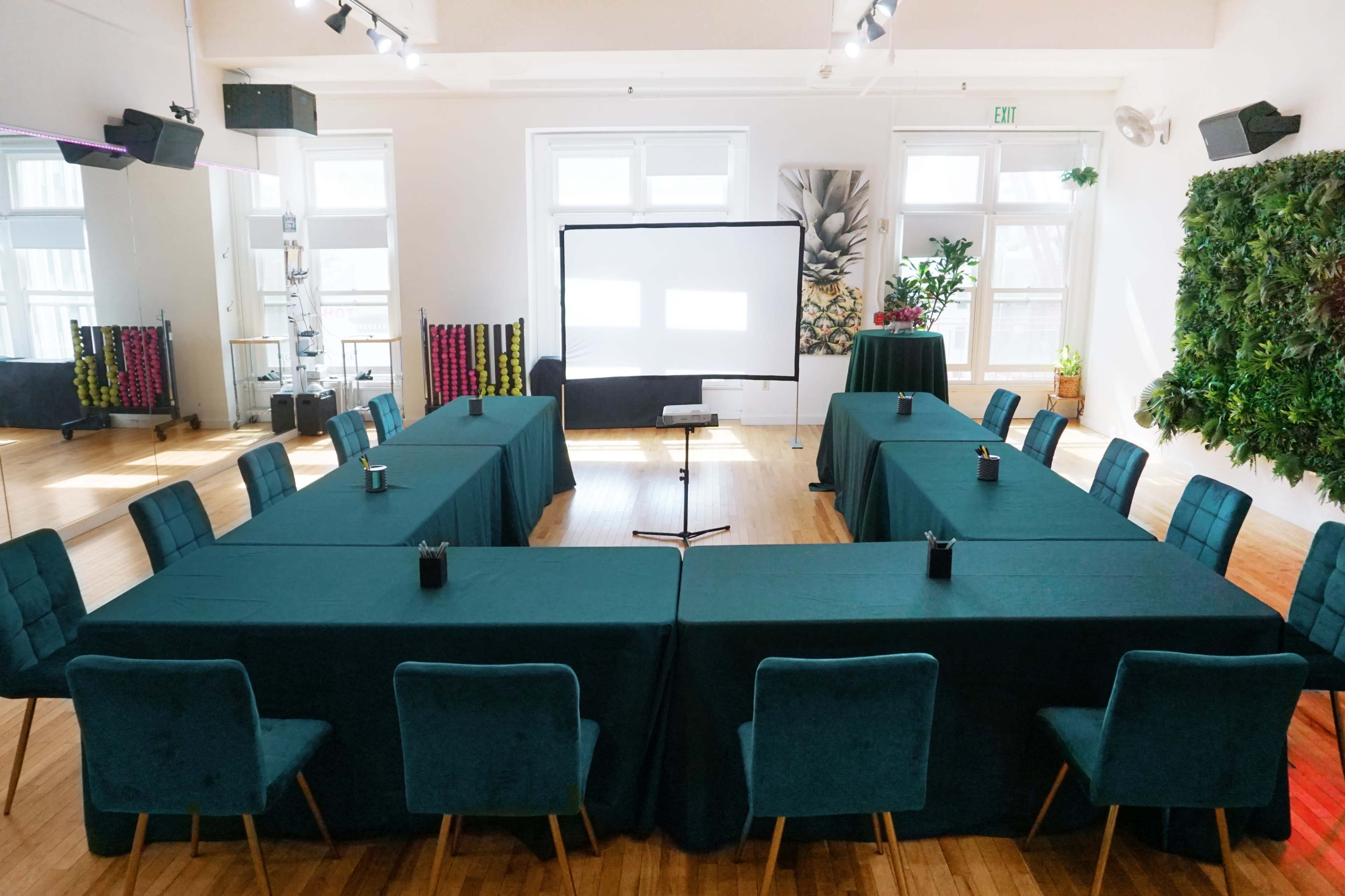 A conference room is set up with a large U-shaped arrangement of tables covered in green cloth, facing a screen at the front of the room.