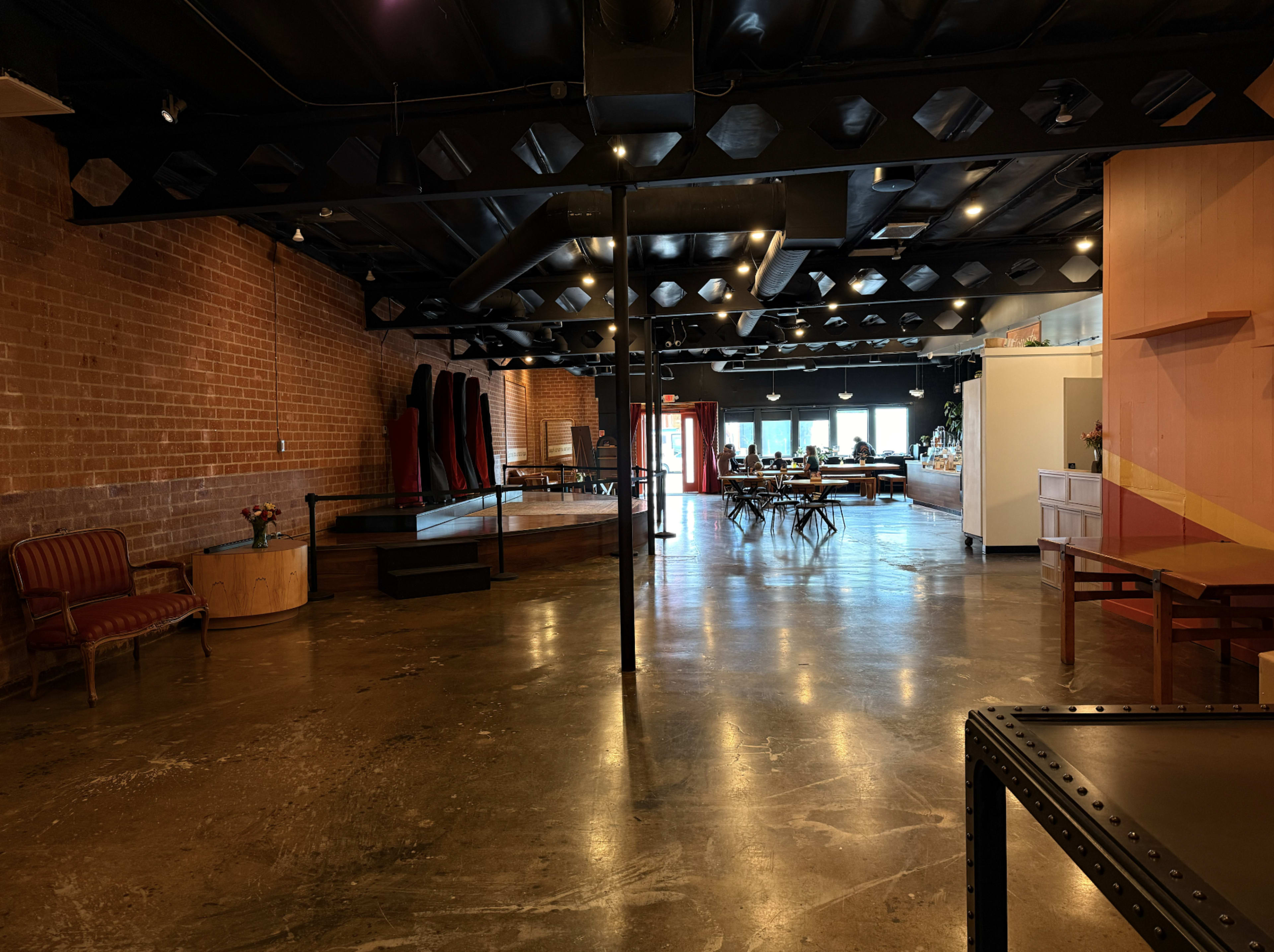 The image shows an expansive, modern interior space with a polished floor, exposed brick walls, various seating arrangements, and distinct areas for gatherings and work.