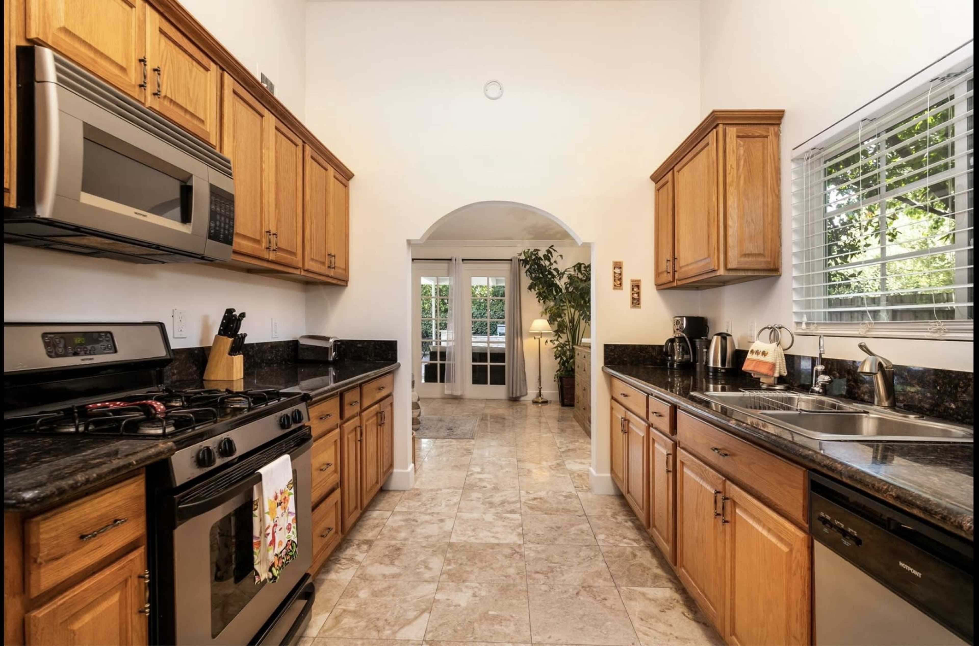 A modern kitchen features wooden cabinets, stainless steel appliances, and a tiled floor, leading to an archway that opens into a well-lit area with plants and a lamp.