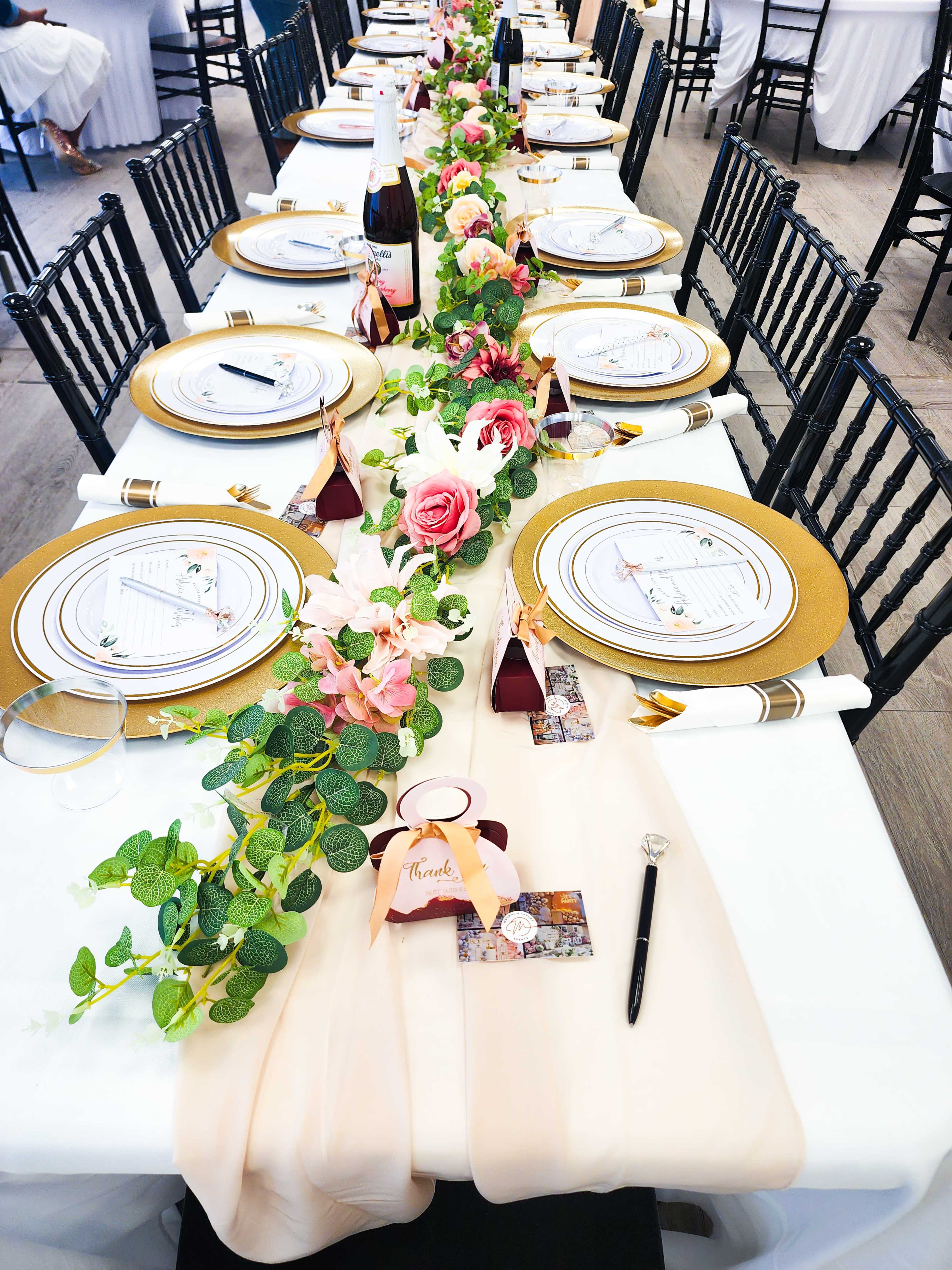 A long banquet table is elegantly set with gold-rimmed plates, floral centerpieces, and decorative place cards, flanked by black chairs.