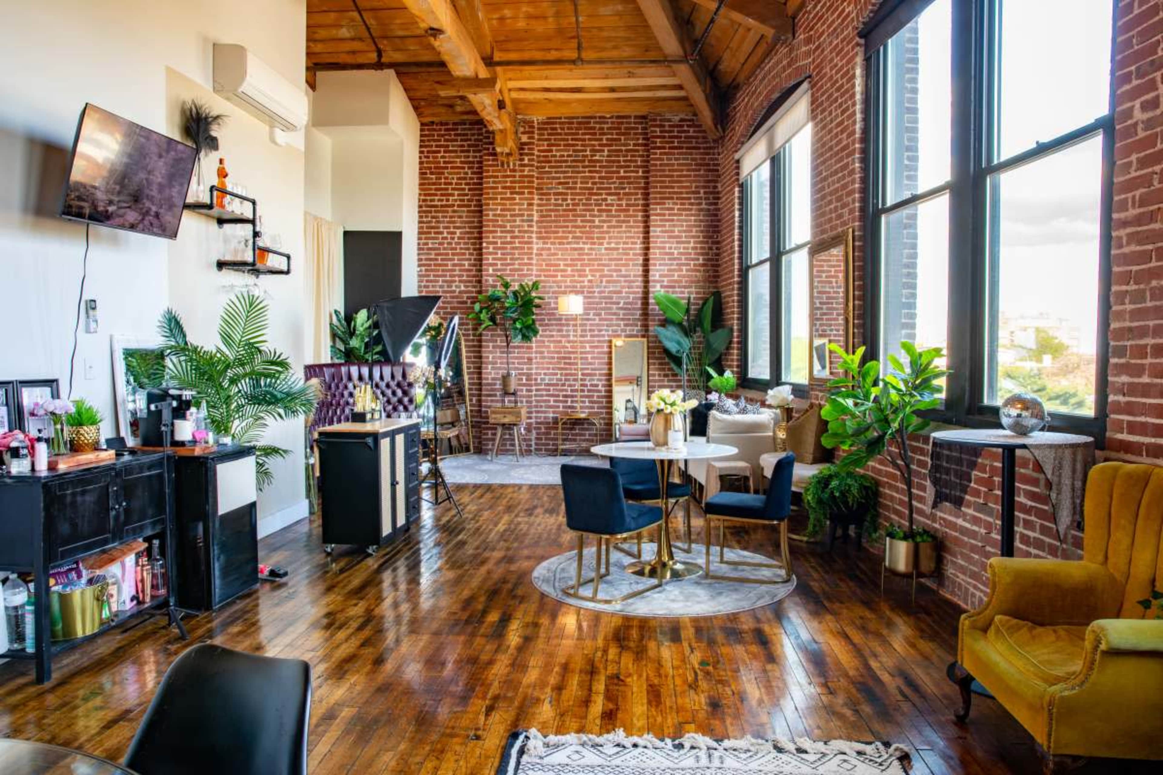 A spacious loft-style room with exposed brick walls, wooden beams, and various plants, featuring a seating area with chairs and a table.