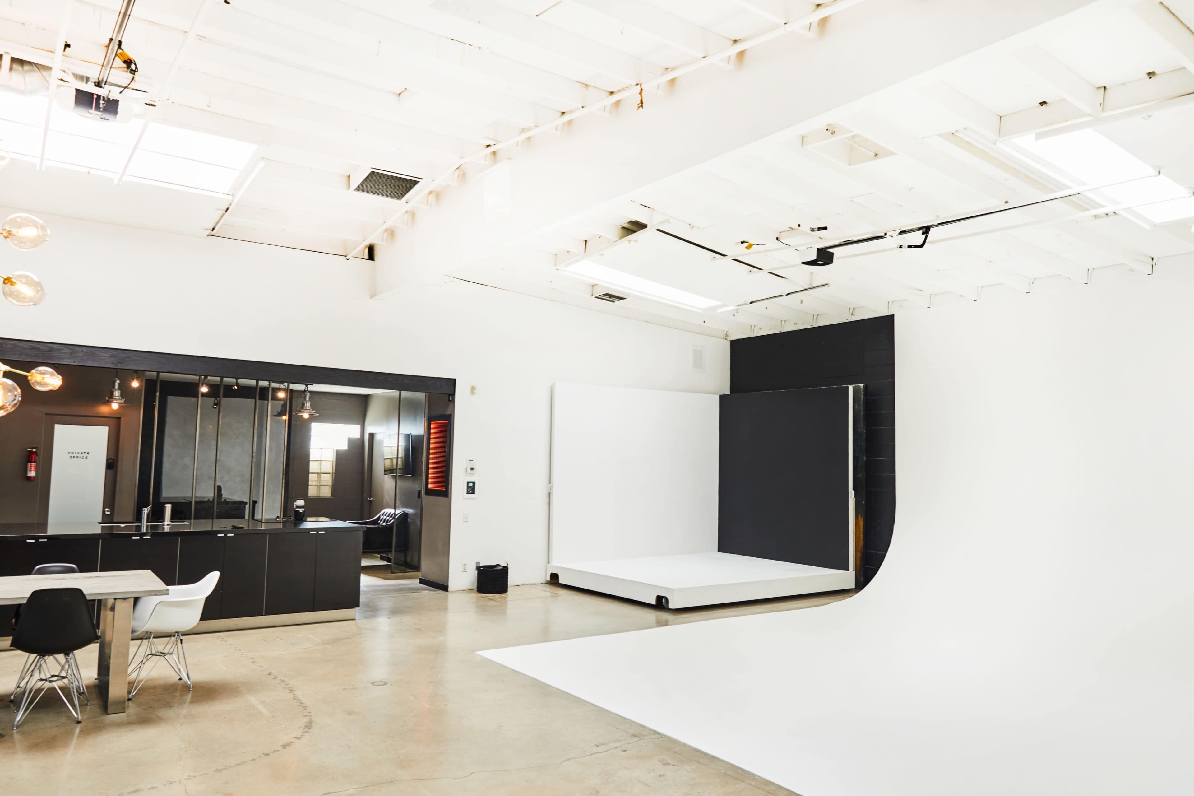 A spacious studio featuring a large white backdrop, polished concrete flooring, a black kitchen area, and modern lighting fixtures.