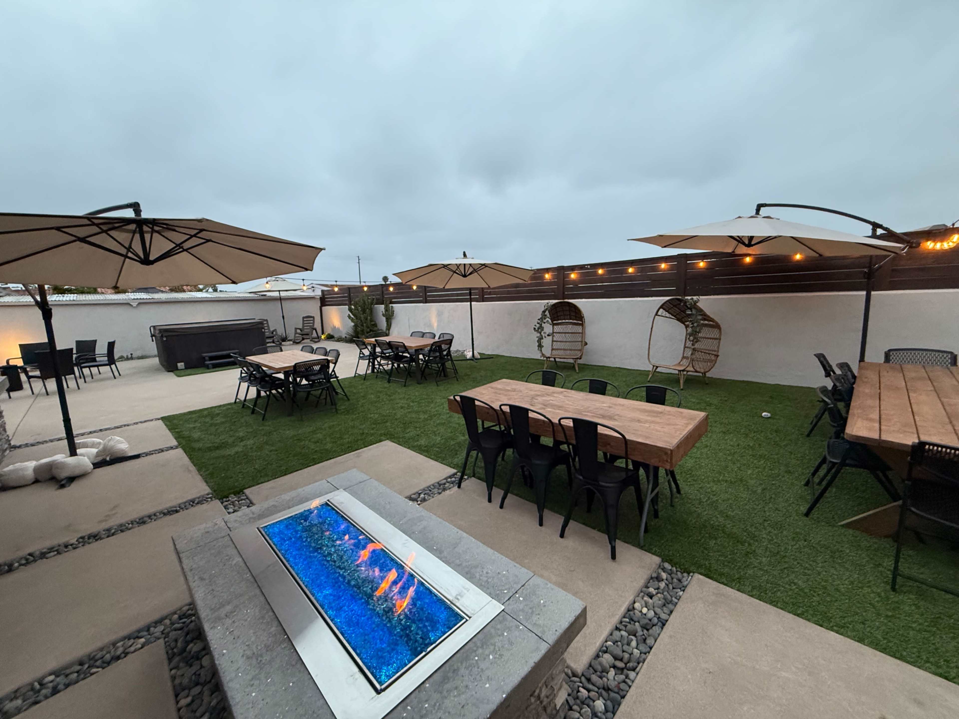 The image shows an outdoor patio area with tables, chairs, umbrellas, and a fire pit surrounded by grass and a wall.