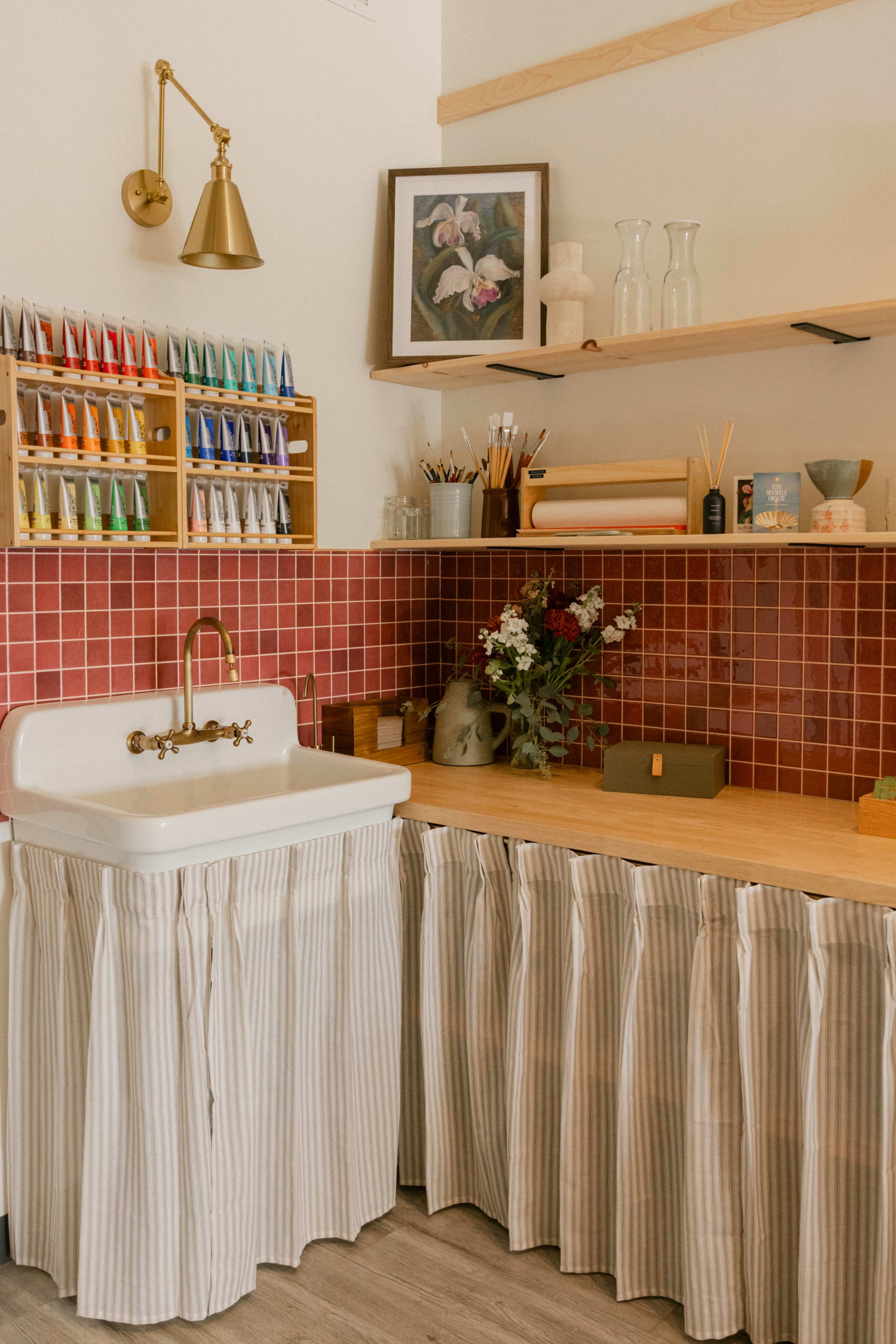 The image shows a cozy art studio with a white sink, wooden shelves displaying paint tubes, and a red tiled backsplash, all complemented by a floral painting and various art supplies.