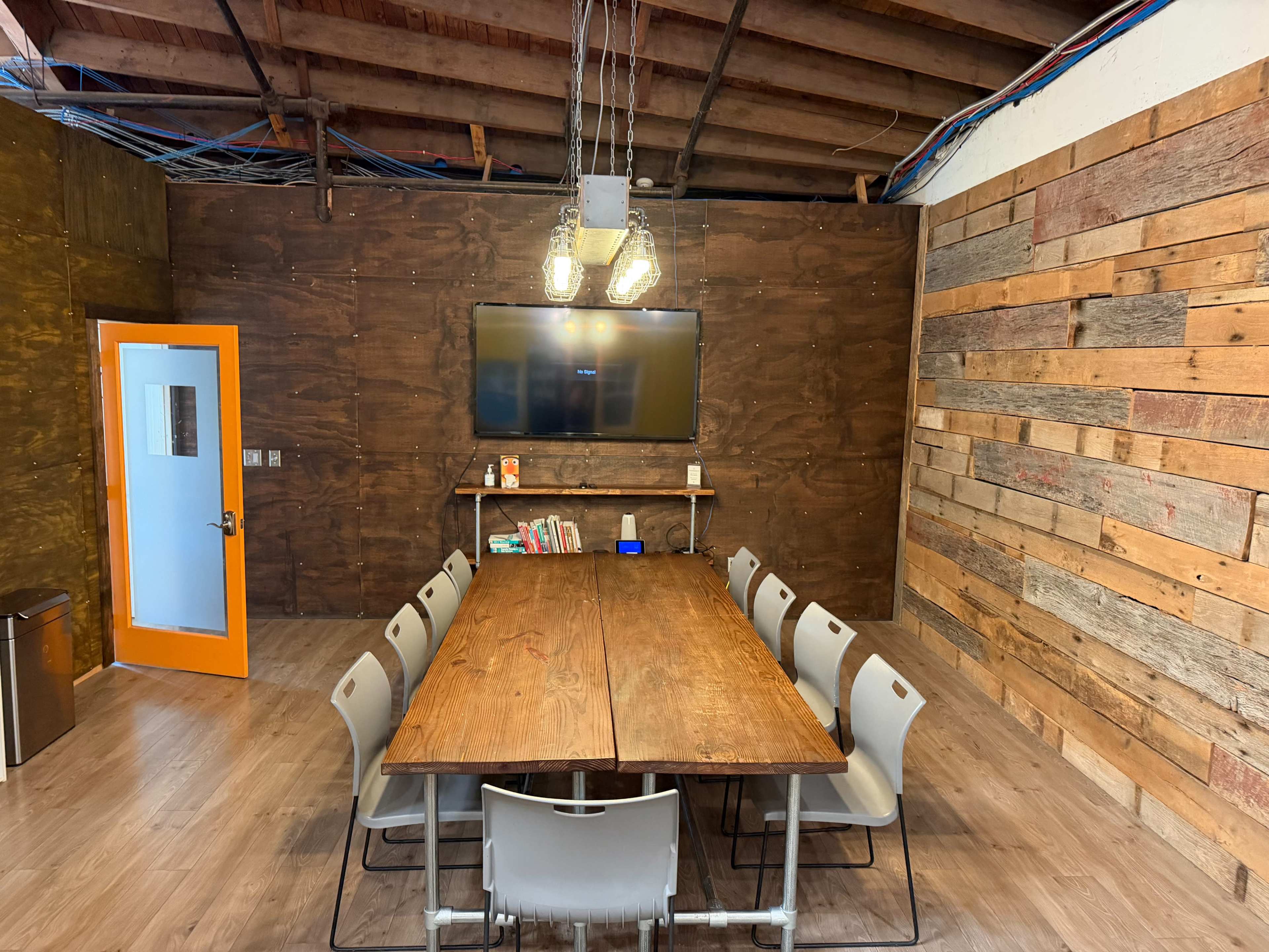 The image shows a conference room with a large wooden table surrounded by ten chairs, a television mounted on the wall, and wooden panels on the walls, along with an orange door.