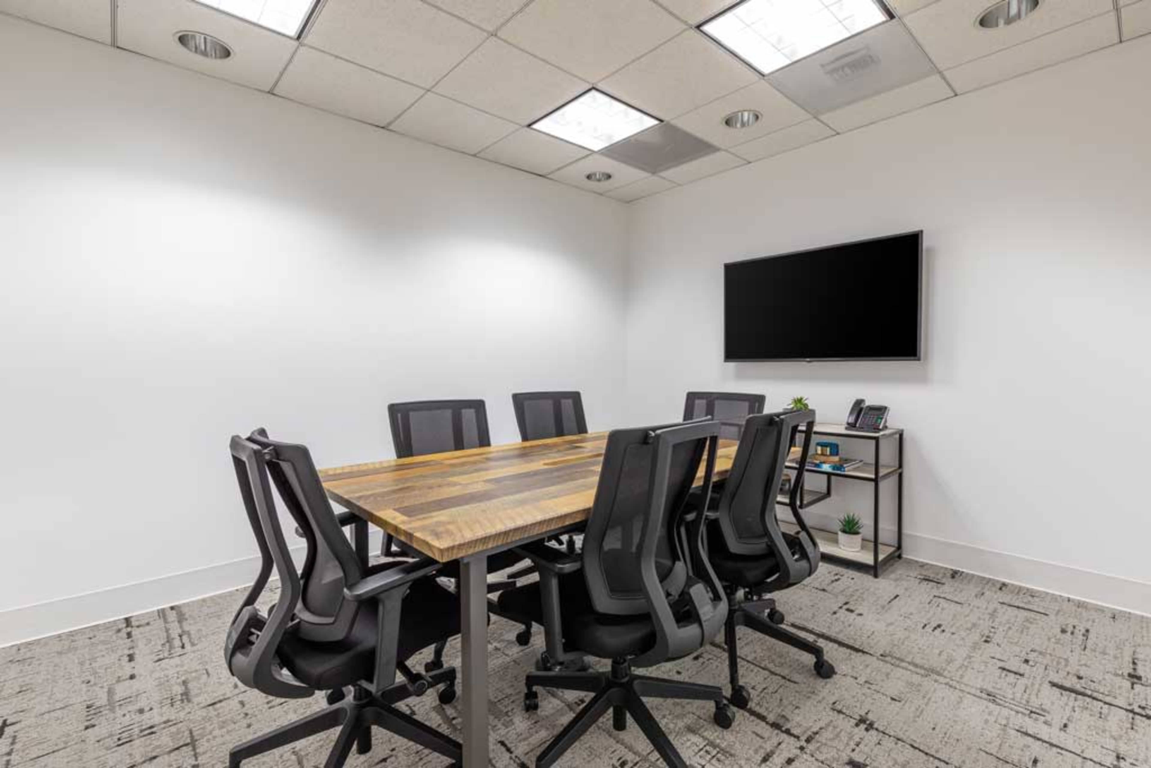 A modern conference room features a wooden table surrounded by six black office chairs, with a television mounted on the wall and a small shelf holding plants and office supplies.