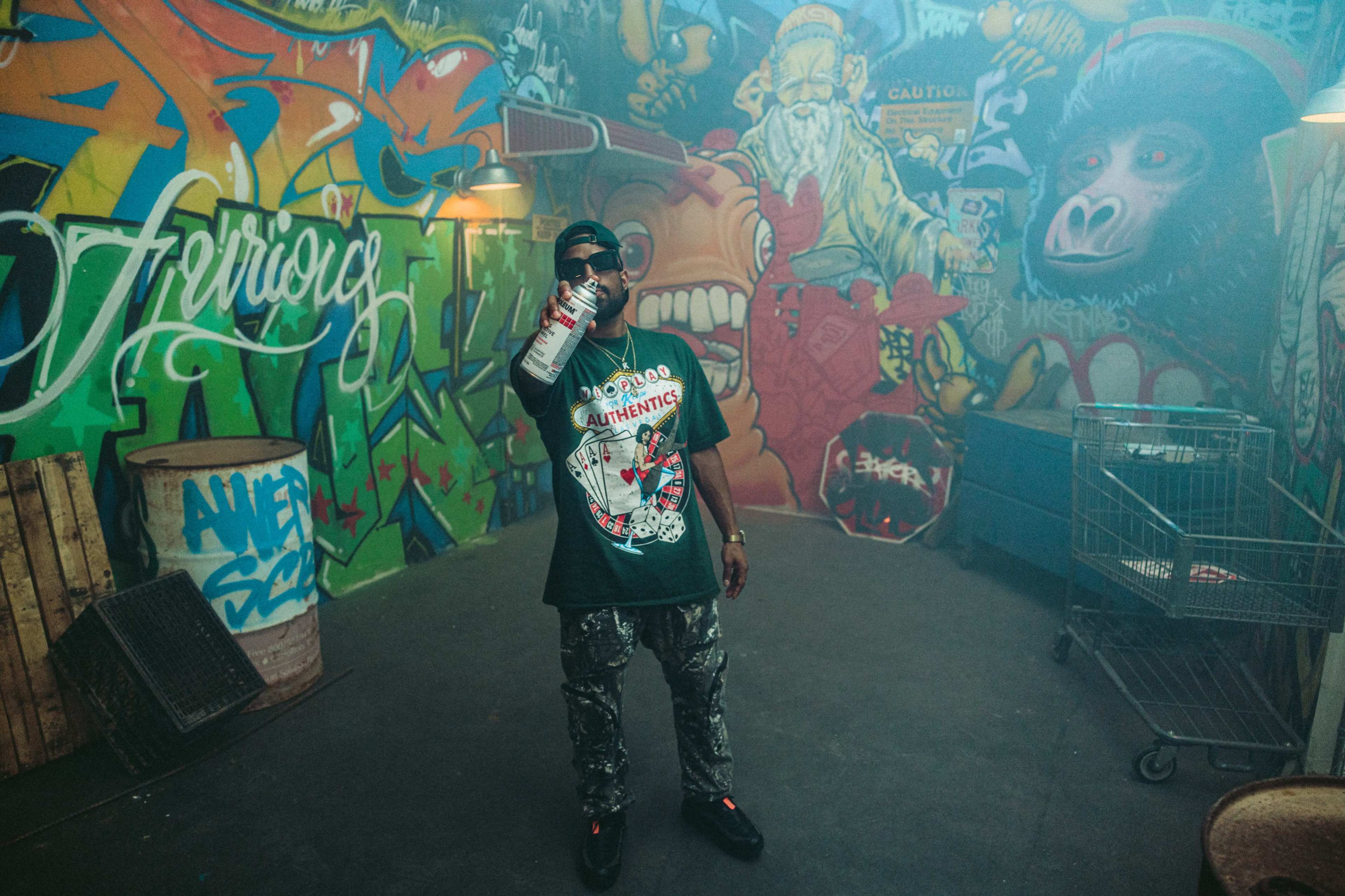 A person stands in a graffiti-covered room, holding a spray can and wearing a graphic t-shirt.