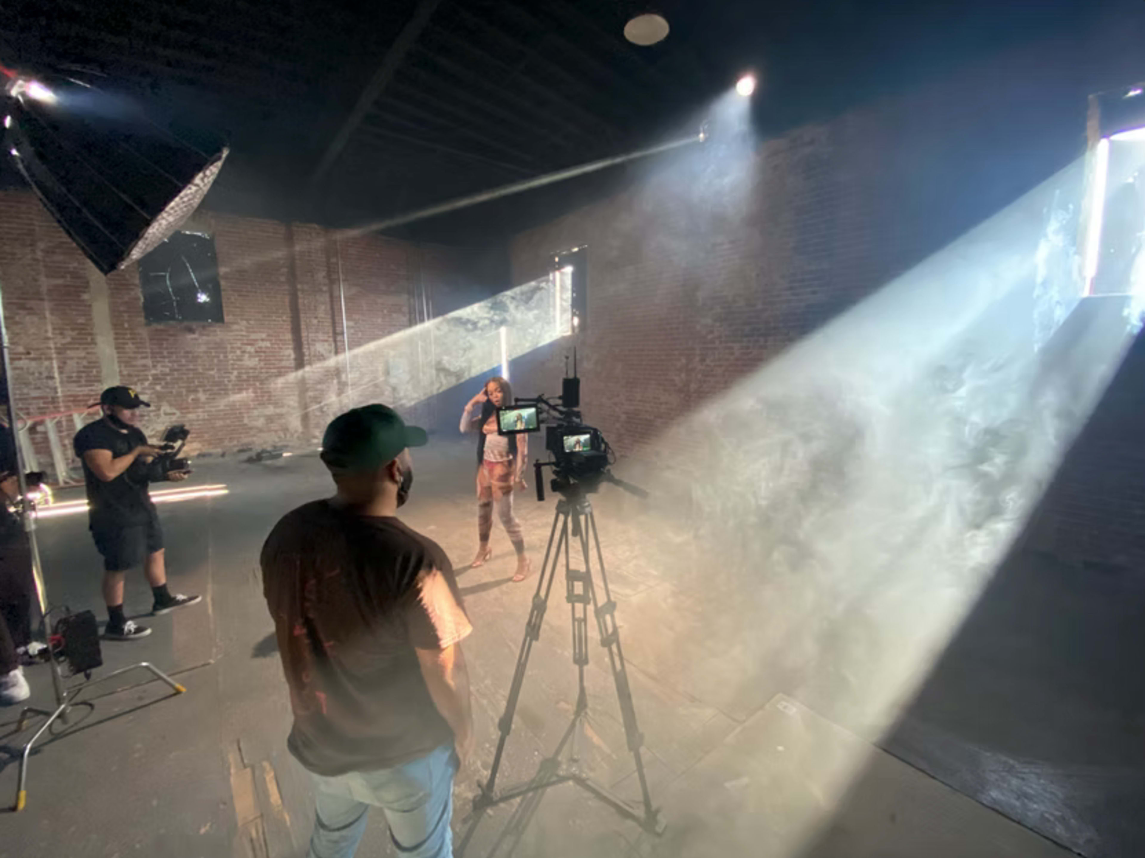 A male model poses in front of a camera while two crew members operate equipment in a dimly lit studio with rays of light streaming through the window.