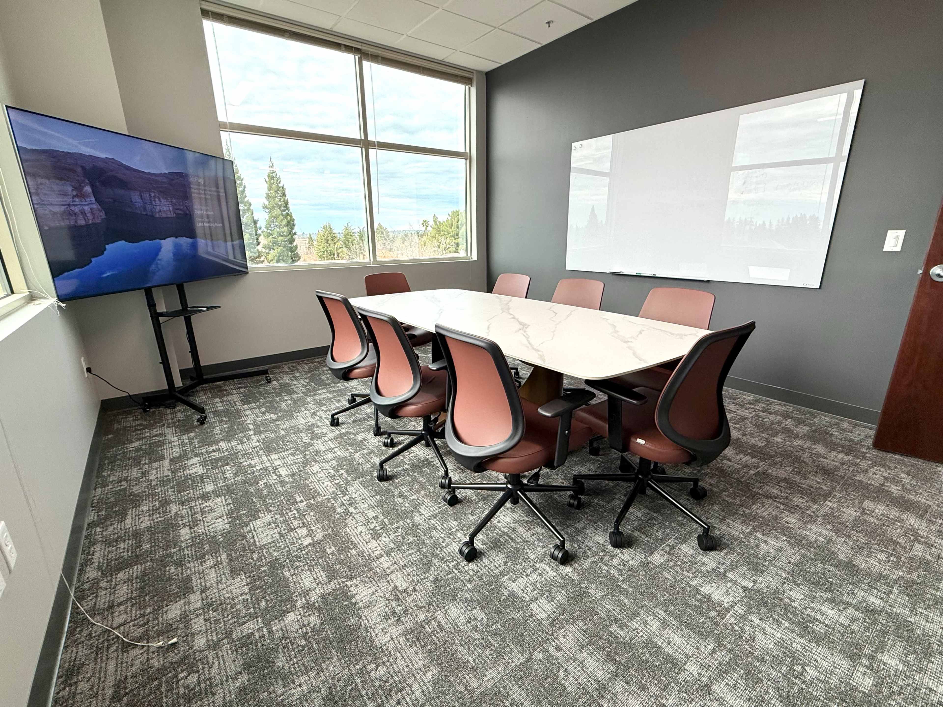 The image shows a modern meeting room featuring a long table surrounded by eight chairs, a large screen, and a whiteboard, with windows providing a view of trees outside.