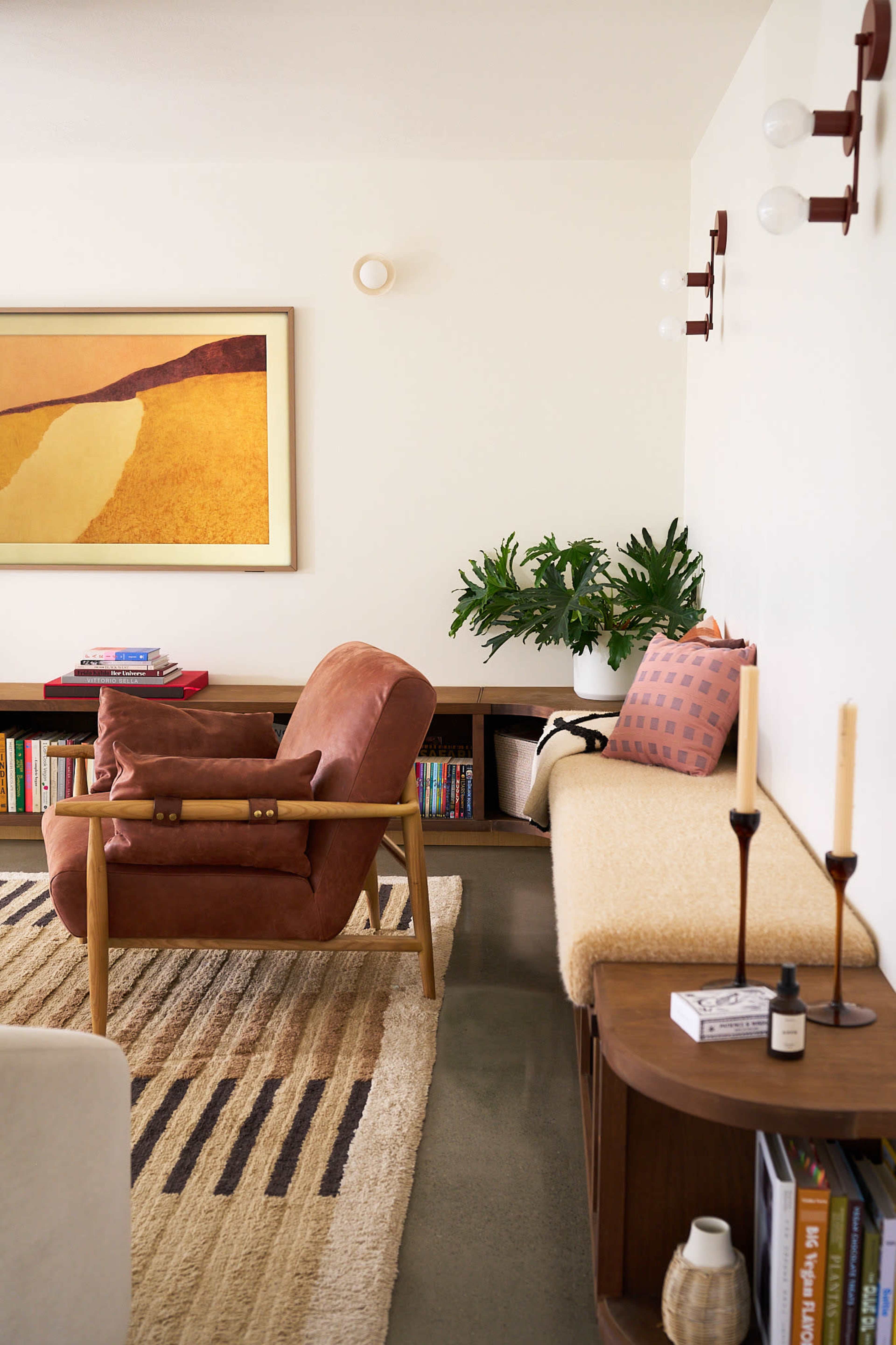 The image shows a cozy living room featuring a brown upholstered chair, a long bench with a patterned cushion, a small wooden table, and a wall adorned with modern lighting and artwork.