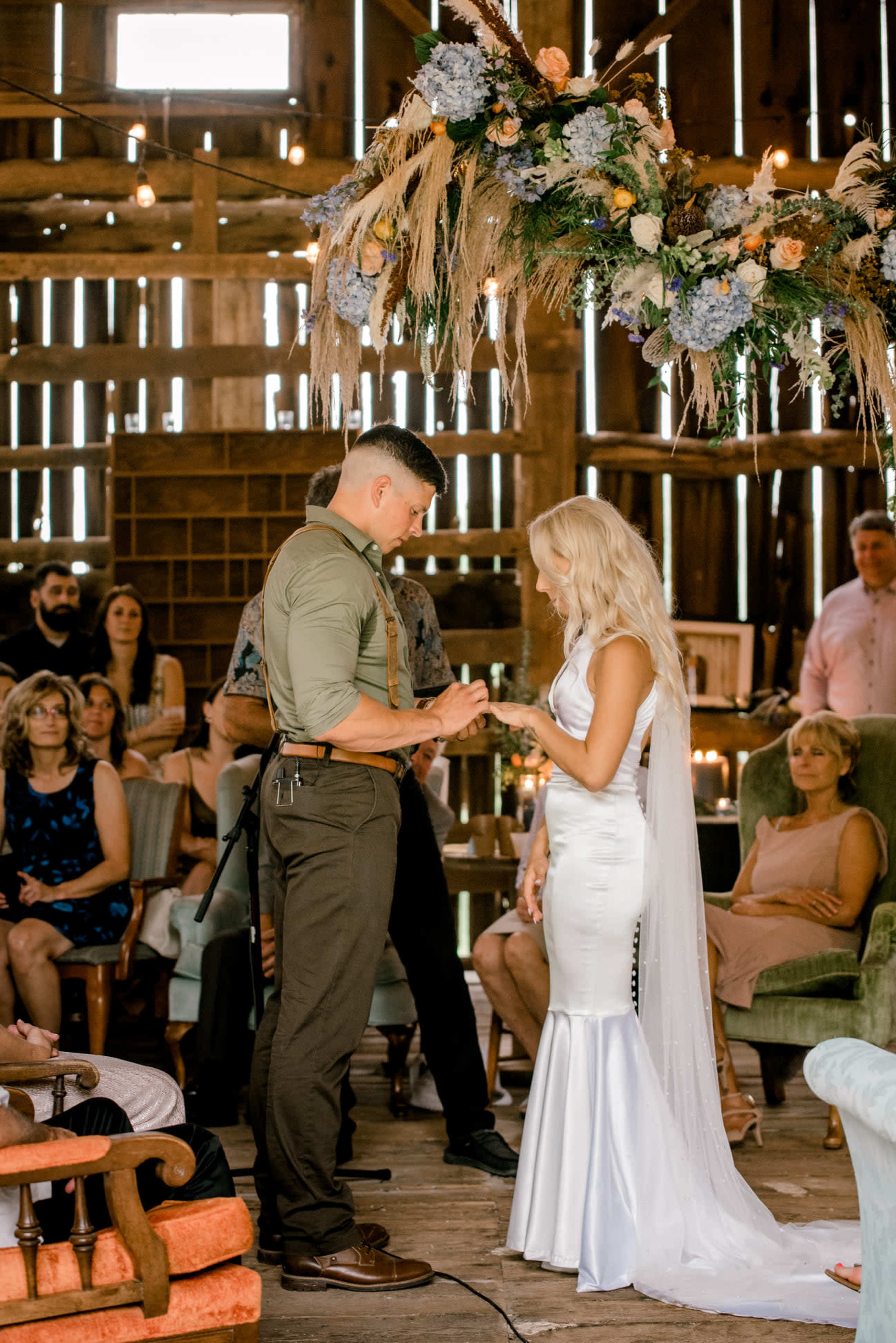 A couple exchanges vows in a rustic barn decorated with flowers while guests watch in the background.